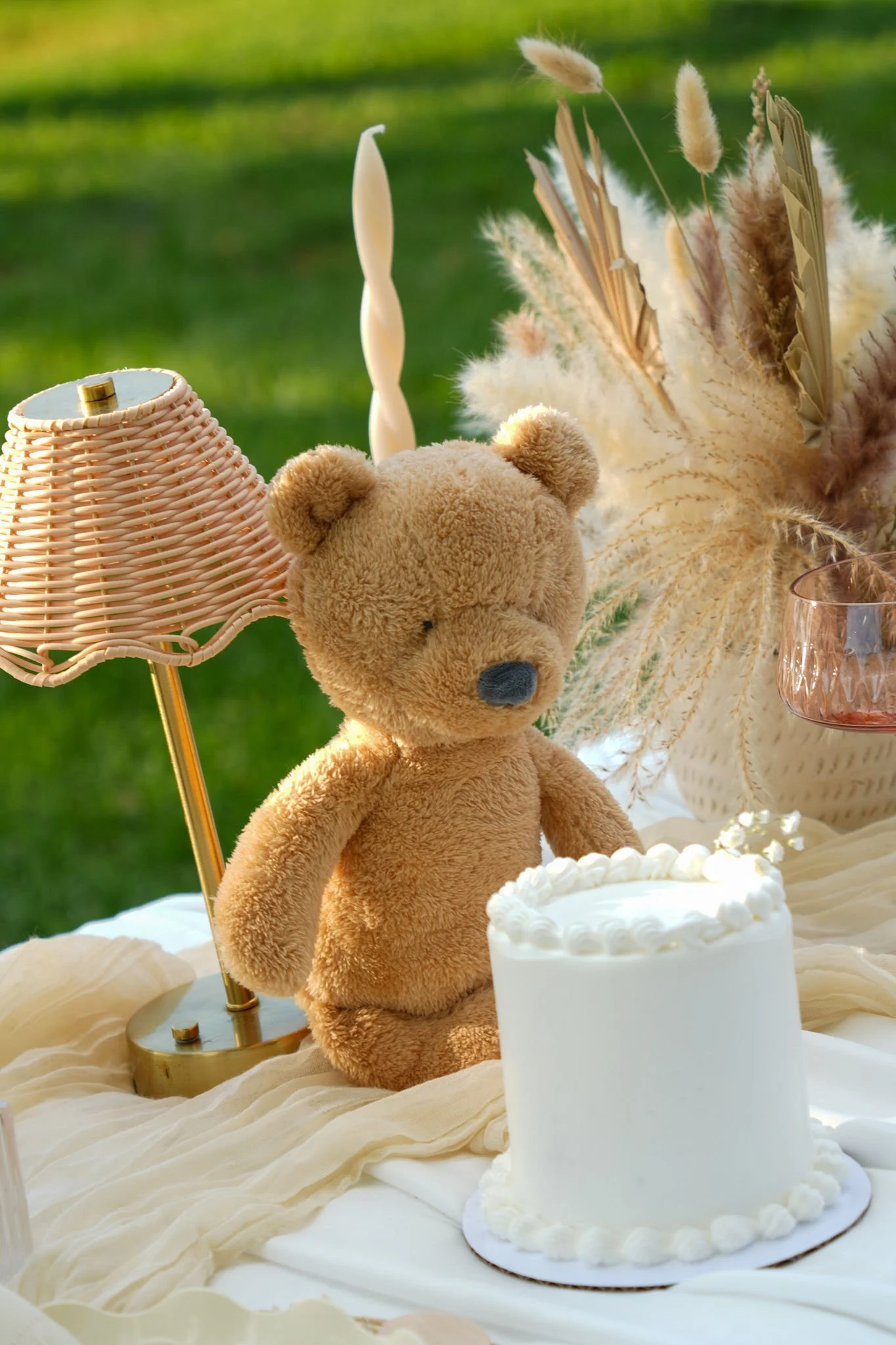 A teddy bear, a white cake with decorative piping, a lamp with a ruffled shade, and decorative dried grasses on a table set outdoors with a green background.