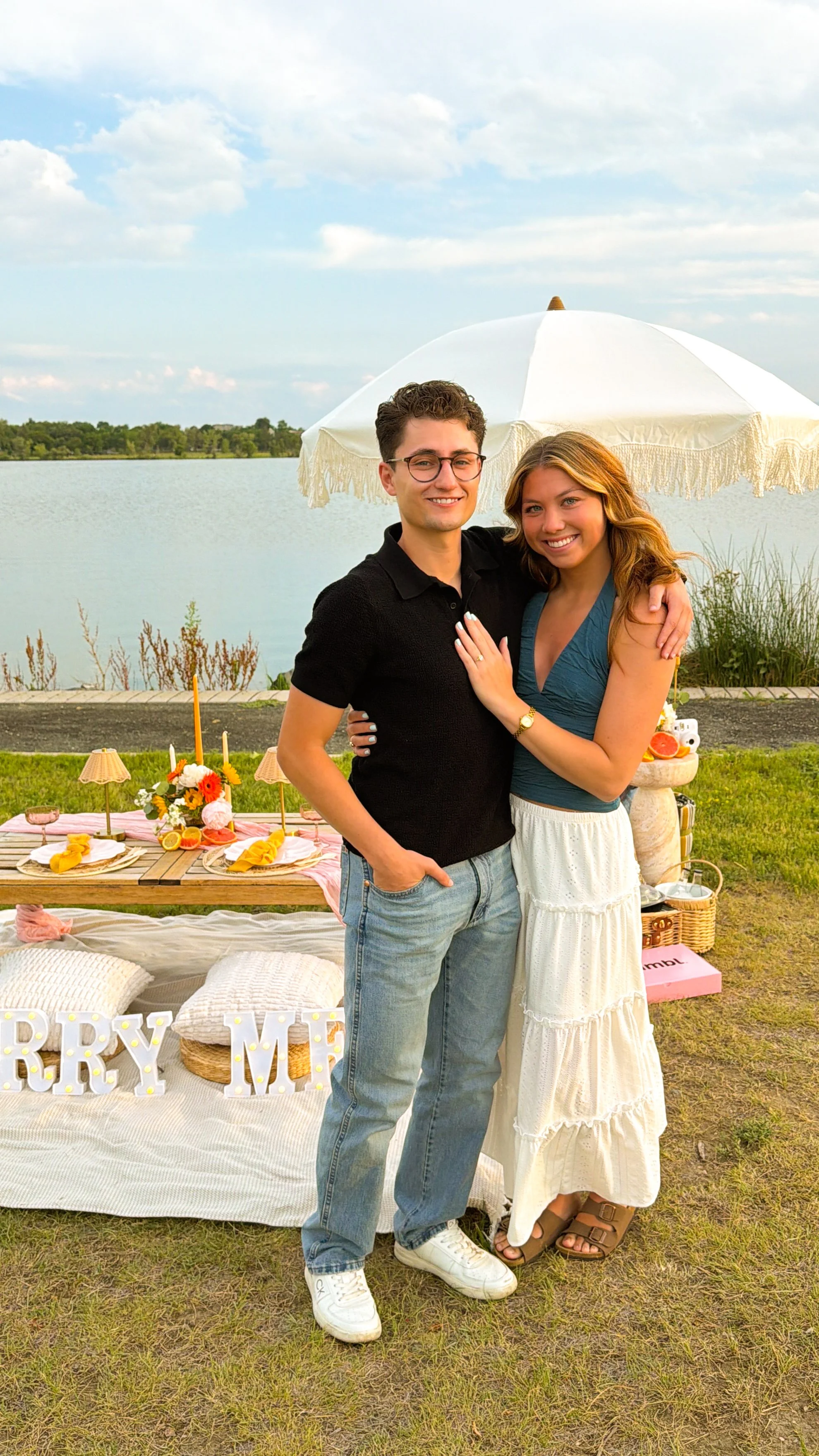 A young couple standing close together outdoors by a lake, smiling at the camera. They are surrounded by a decorated picnic setup with a white umbrella, flowers, candles, and a table set for a celebration, with decorative pillows.