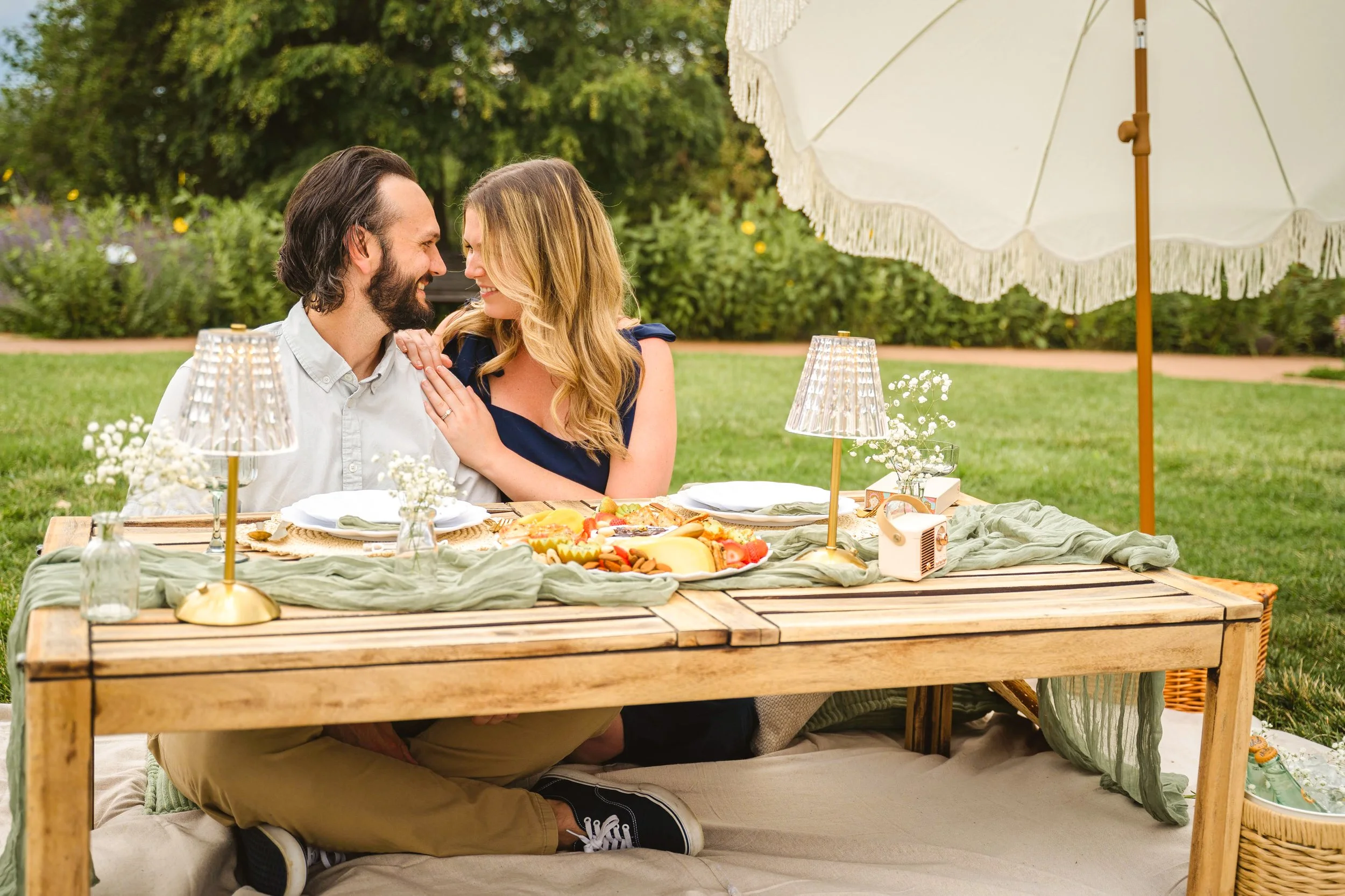 A couple sitting at a picnic table outdoors under a large white patio umbrella, sharing a happy moment, with food and drinks on the table.