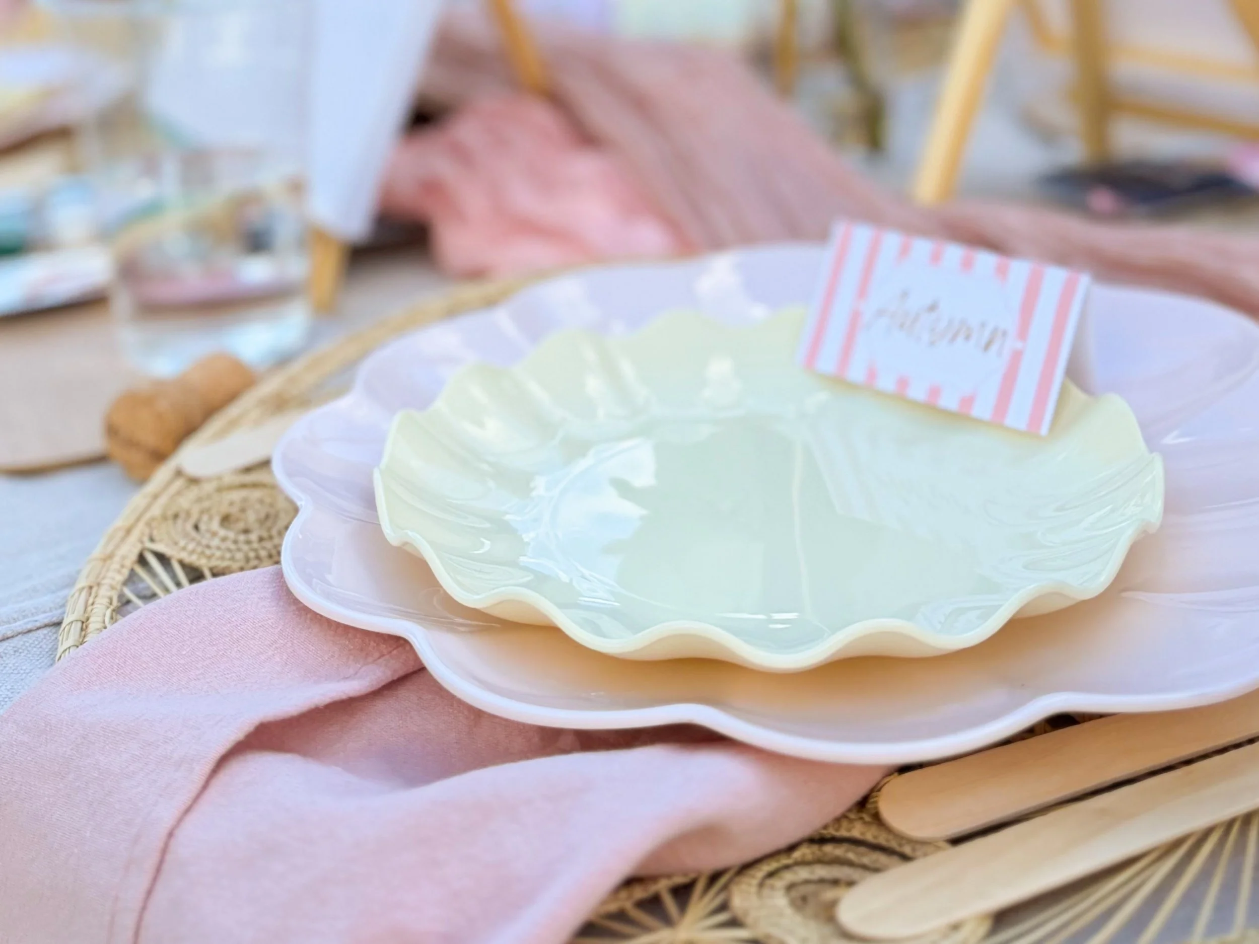 A table setting for a luxury picnic in Denver, Colorado, showing pink and beige scalloped plates, bamboo cutlery, pink napkin, and place card.