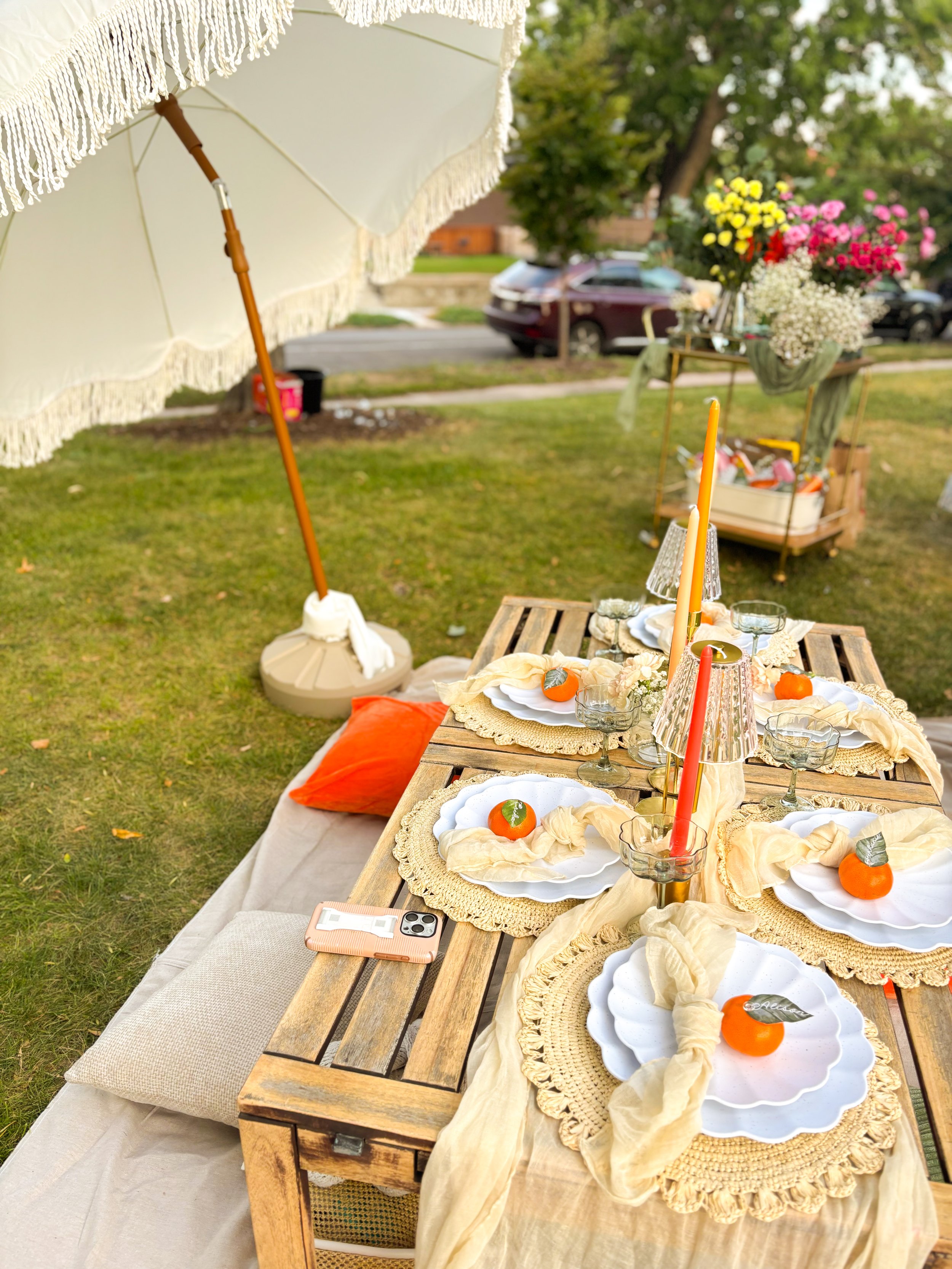 Outdoor tables set with white plates, orange candles, glassware, and decorative tablecloths at a garden party with a large white umbrella and a floral background.