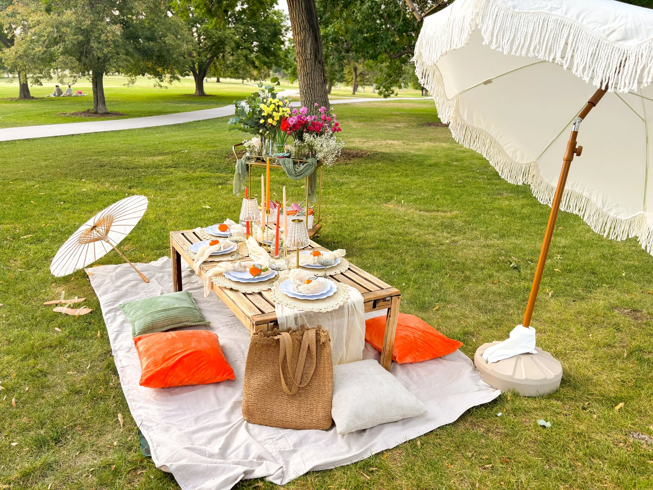 A picnic setup on a blanket in a park with a low table holding flowers, candles, and tableware, surrounded by pillows, with umbrellas providing shade and trees in the background.