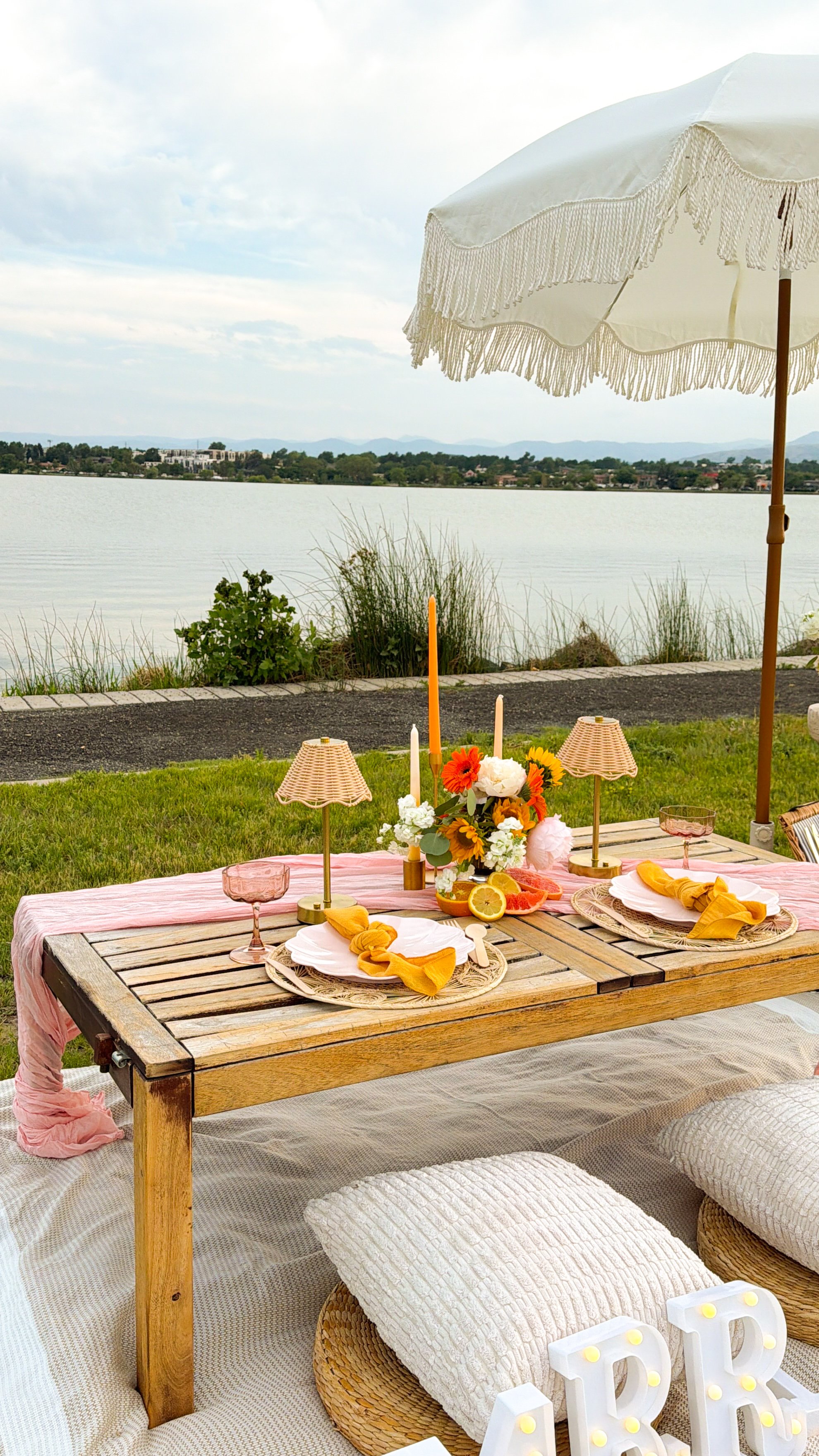 Outdoor dining table setup by a lake with a white umbrella, floral centerpiece, candles, plates, napkins, and decorative items, with mountains and cloudy sky in the background.