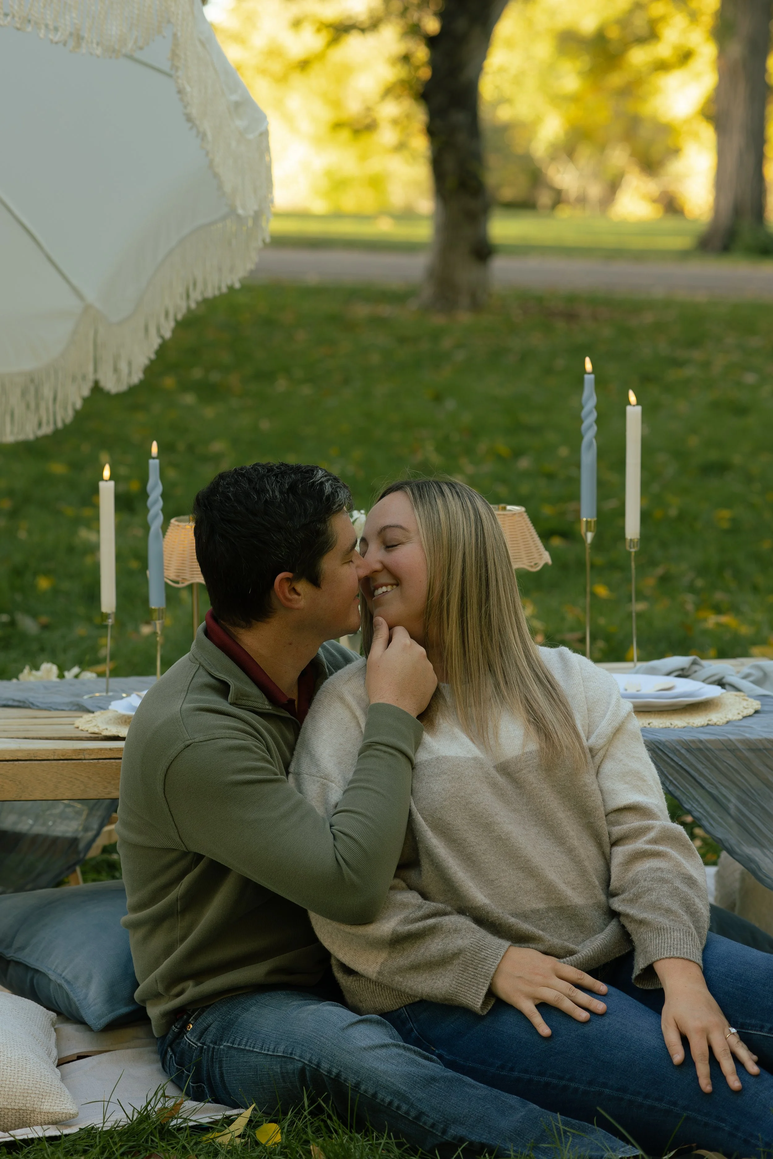 A couple sitting closely together on a picnic blanket outdoors, about to kiss, with candles and a white umbrella in the background, surrounded by trees and autumn foliage.