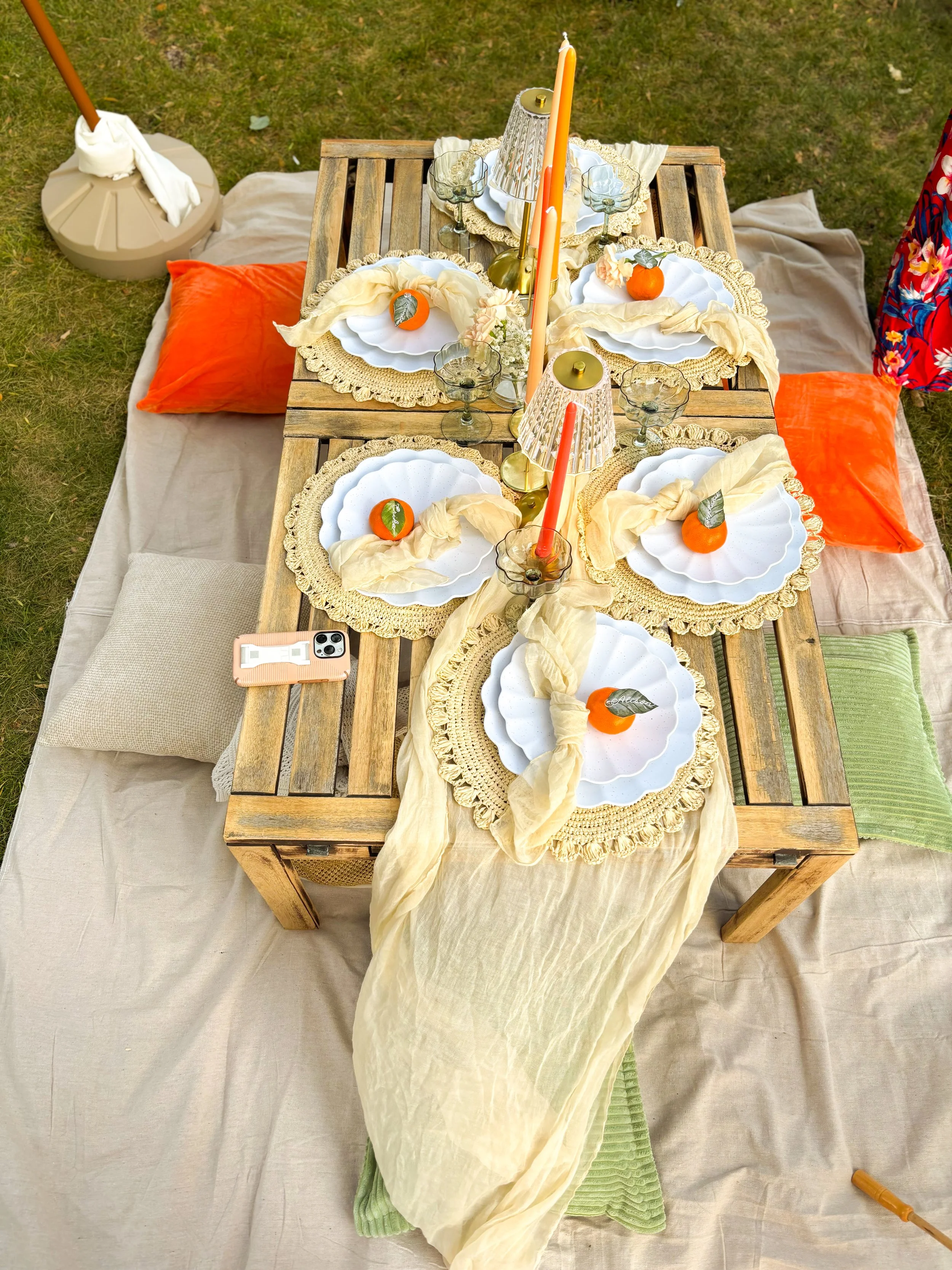 A table set for a picnic with white plates, orange decorative pumpkins with leaves, glass cups, and beige cloth napkins, placed on woven placemats. The table has a yellowish cloth runner, and is surrounded by cushions and blankets on grass.