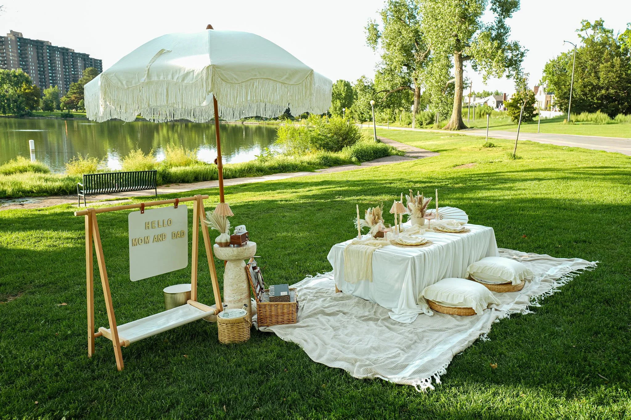 A lakeside outdoor picnic with a white umbrella, a table with a white cloth, decorative candles, and a sign that says 'Hello Mom and Dad.' There is a bench near the water, and trees with a walking path in the background.
