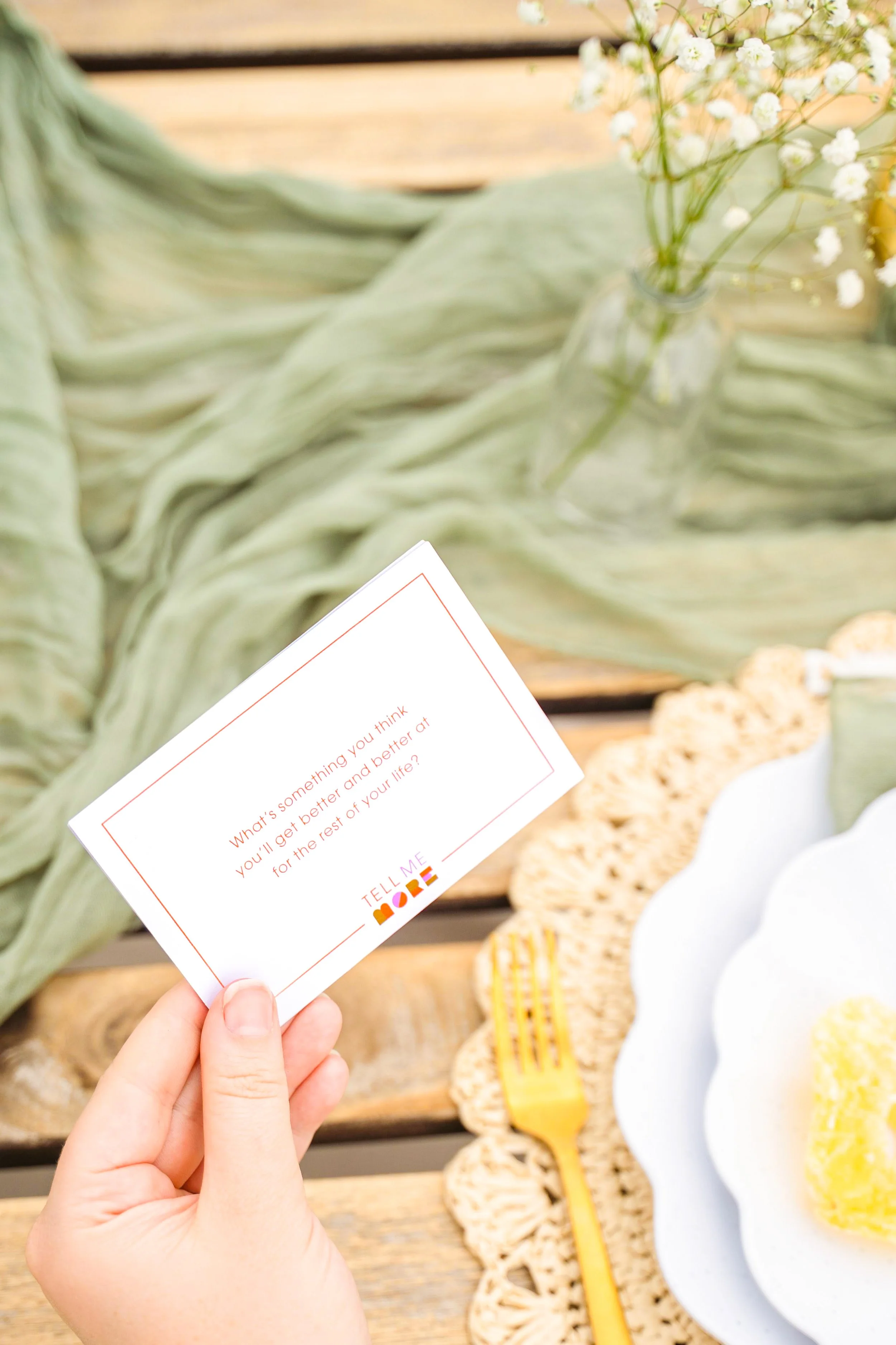 Close-up of a hand holding a small card with a motivational quote, against a background of a wooden table with jars of white flowers and decorative cookies.