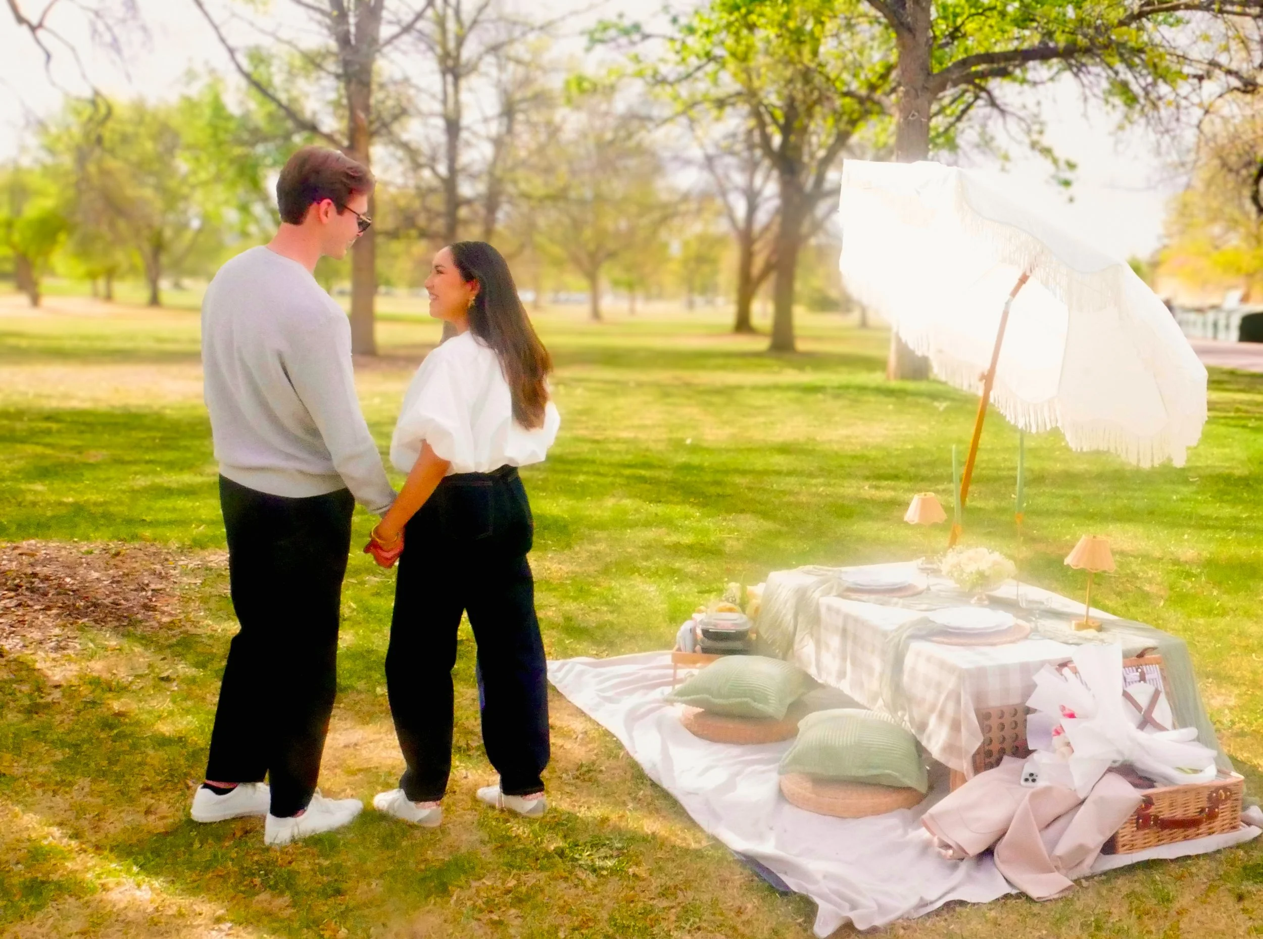 A young man and woman standing in Denver City Park, holding hands and smiling at each other, with a white parasol and a luxury picnic table to the right.