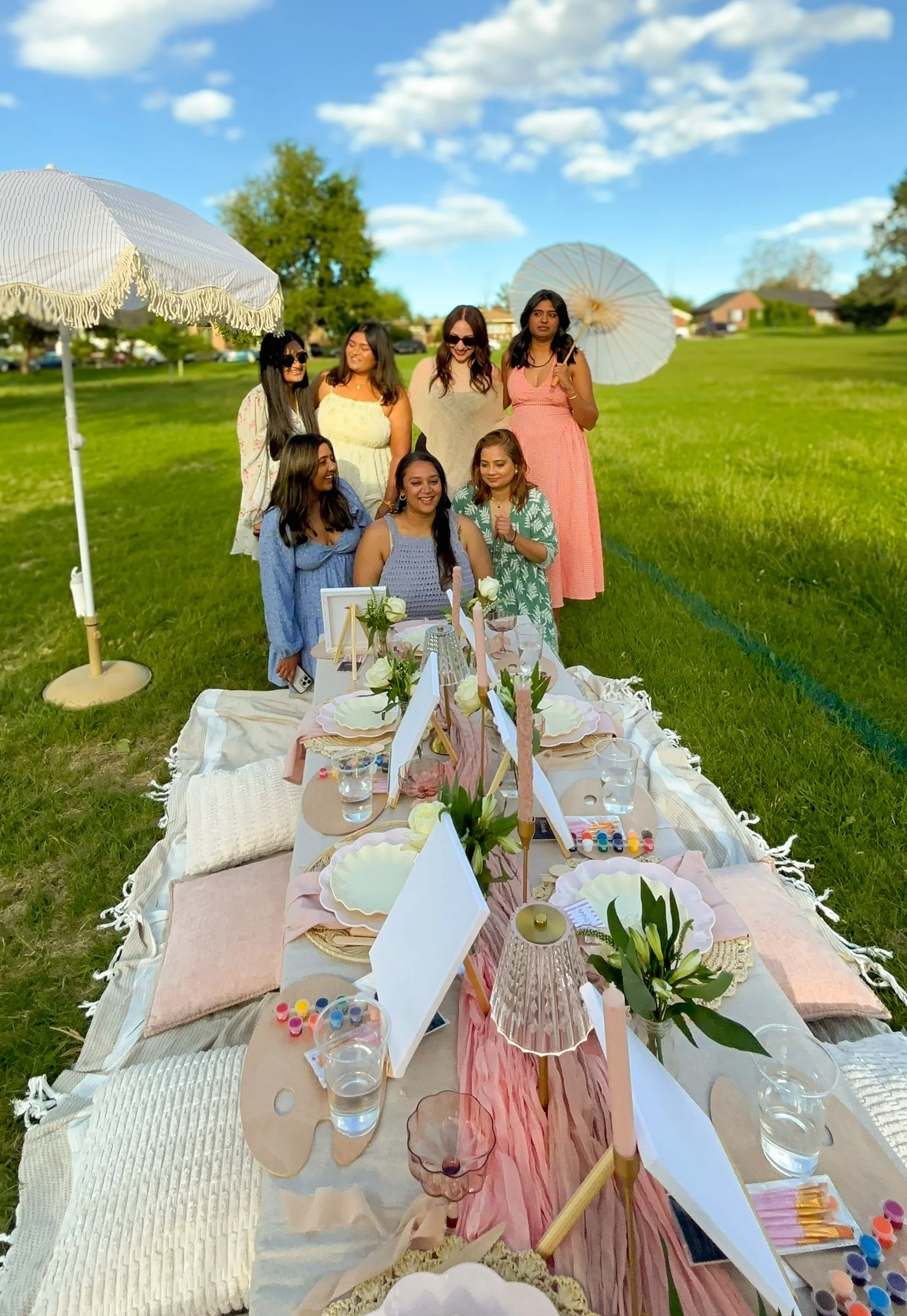 Group of women at a bachelorette luxury picnic with decorated table, umbrellas, and a bright, sunny sky.
