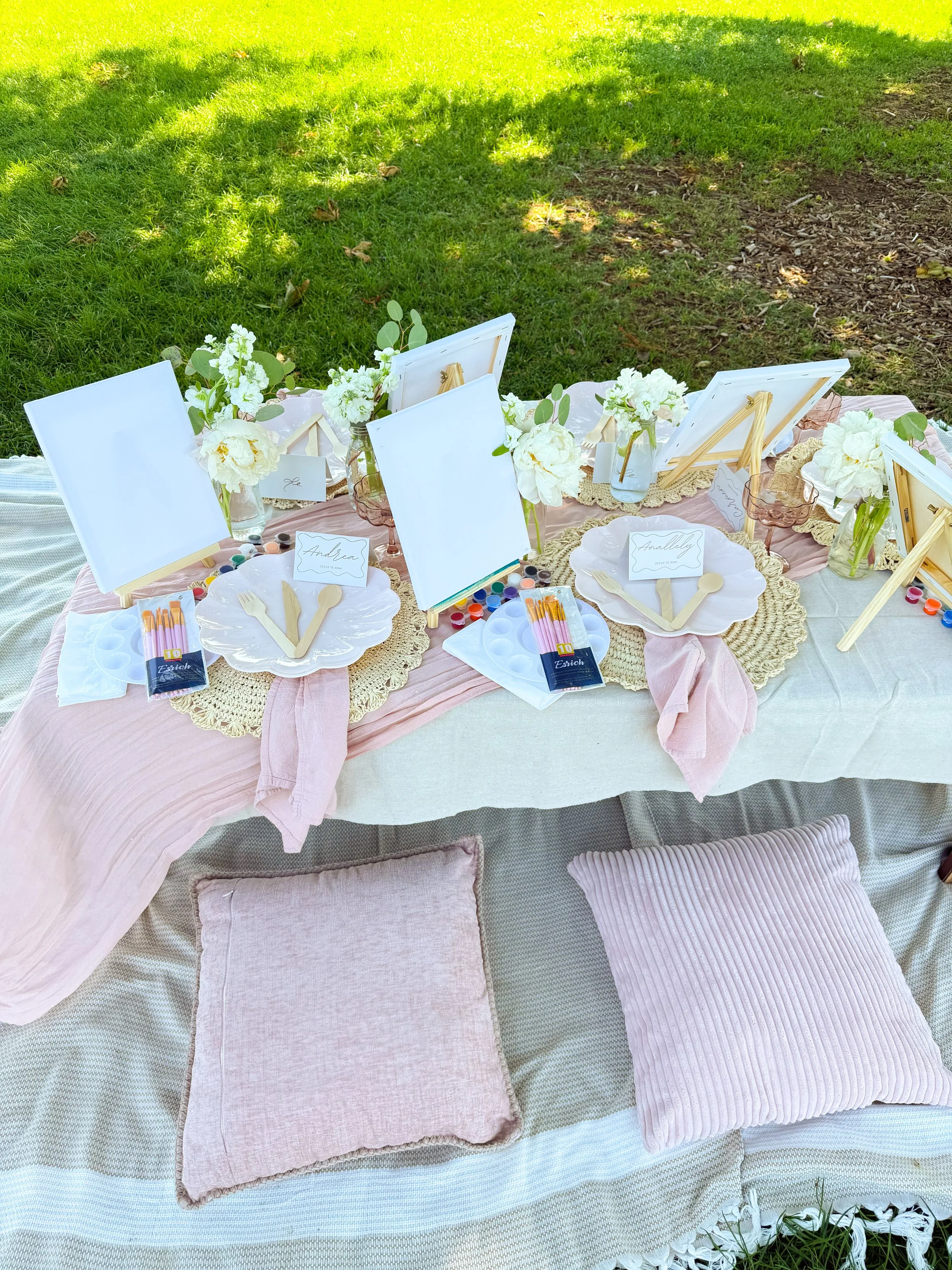 Pink outdoor luxury picnic setup on a blanket with pillows, decorative items, and floral arrangements in a sunny grassy area.