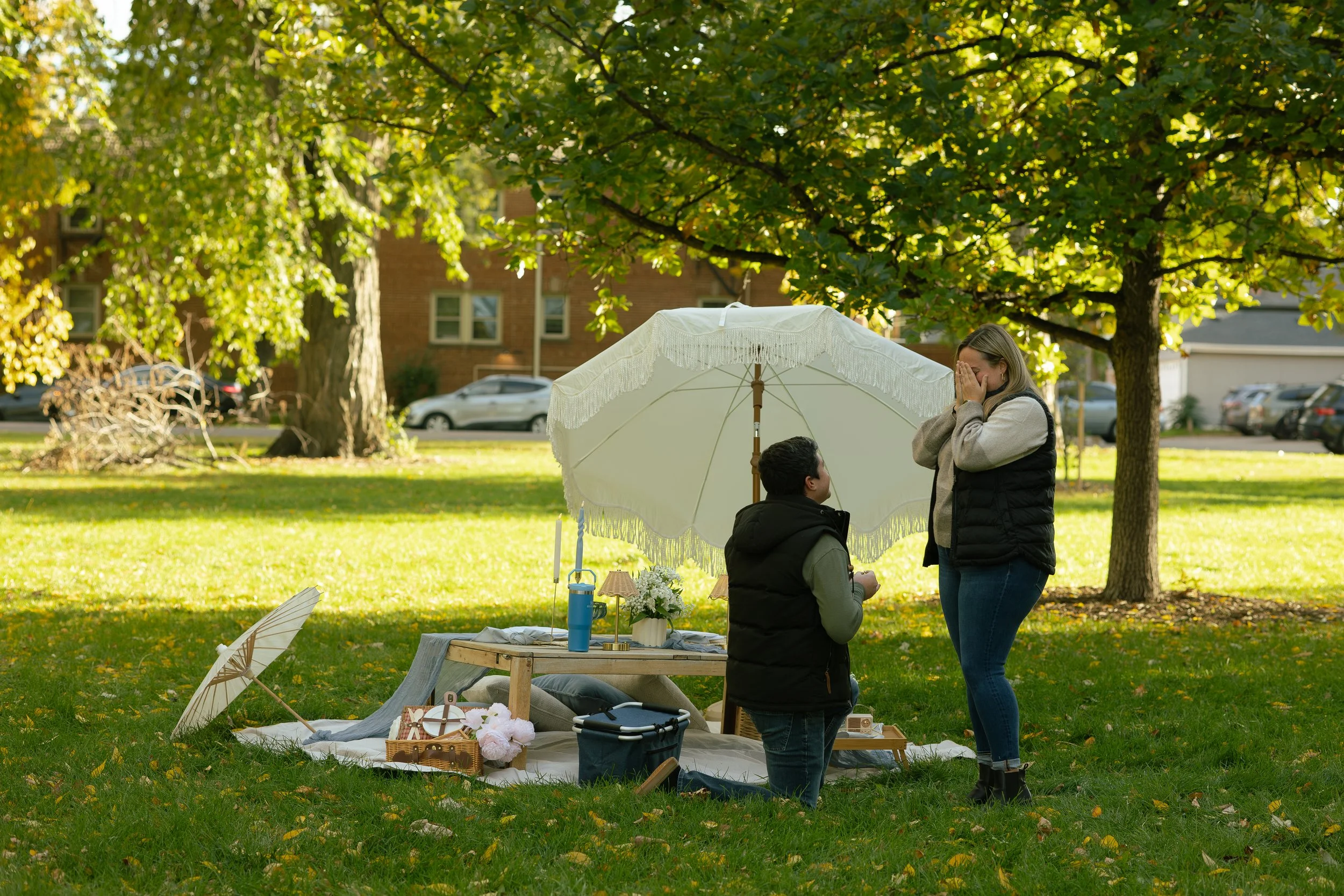 A woman and a man stand under a white parasol on a grassy park area, with a table displaying flowers, and small decorative items. The woman is covering her face with her hands while the man proposes to her.
