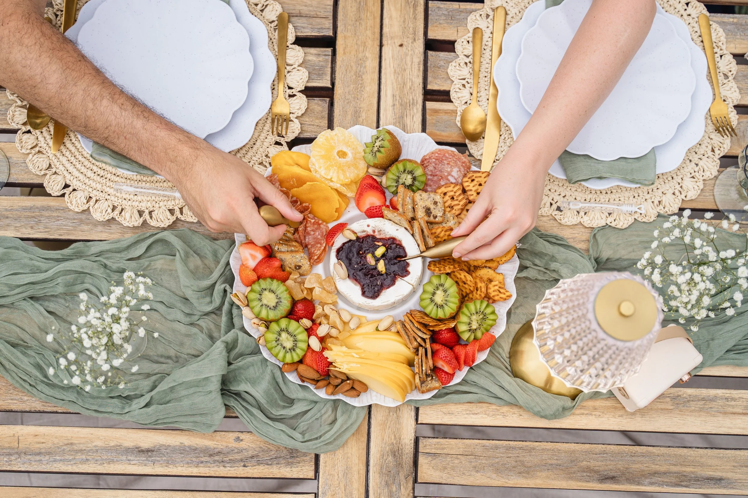 Top view of a table set for a meal with a large platter of assorted fruits, cookies, and a small bowl of jam in the center. The table has woven placemats, white plates, gold utensils, and a green cloth underneath the platter, with some small white fl