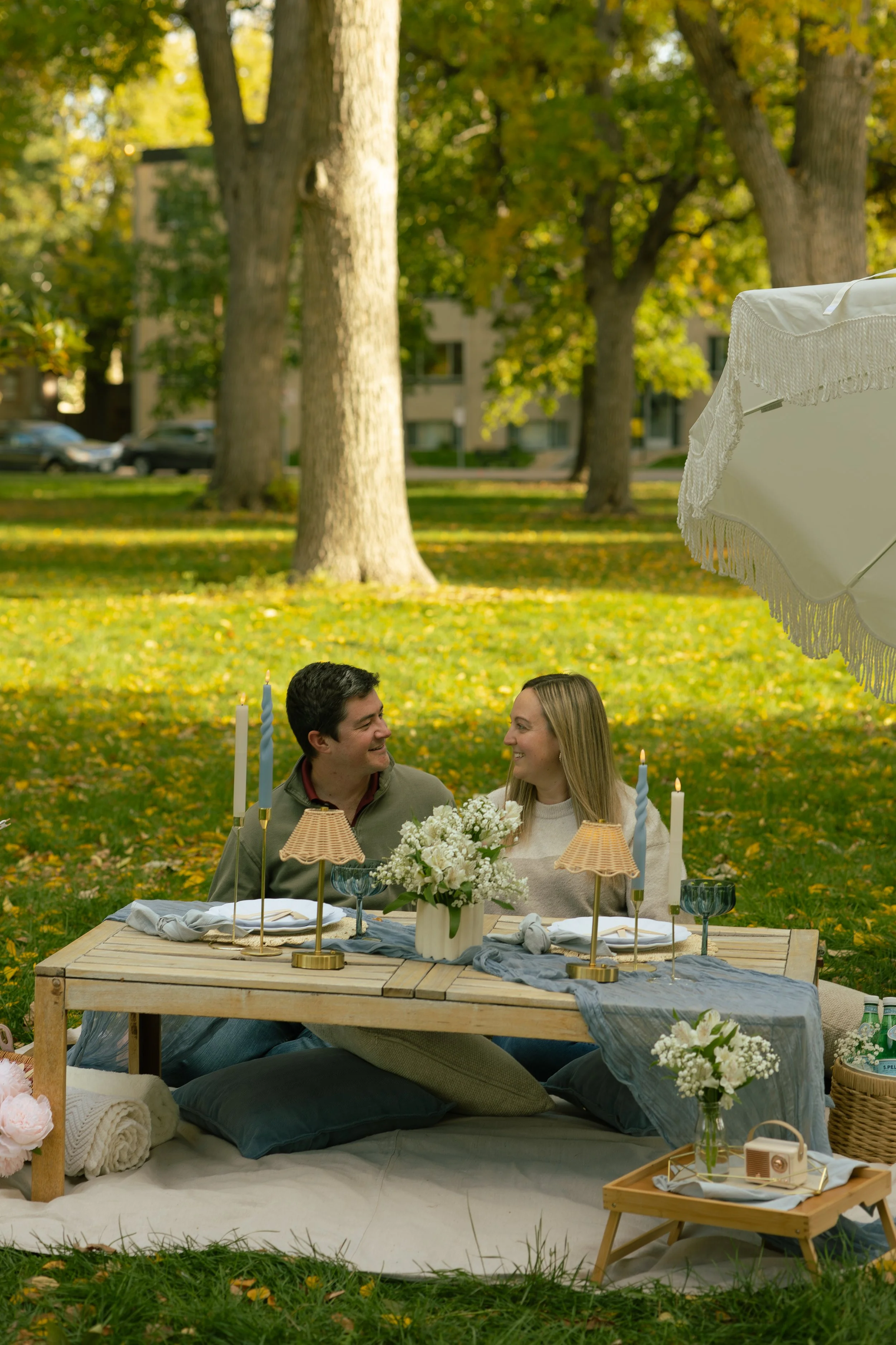 A man and woman sitting at a decorated outdoor table with flowers, candles, and tableware, smiling at each other under a large white umbrella, in a park with green trees and fallen leaves.