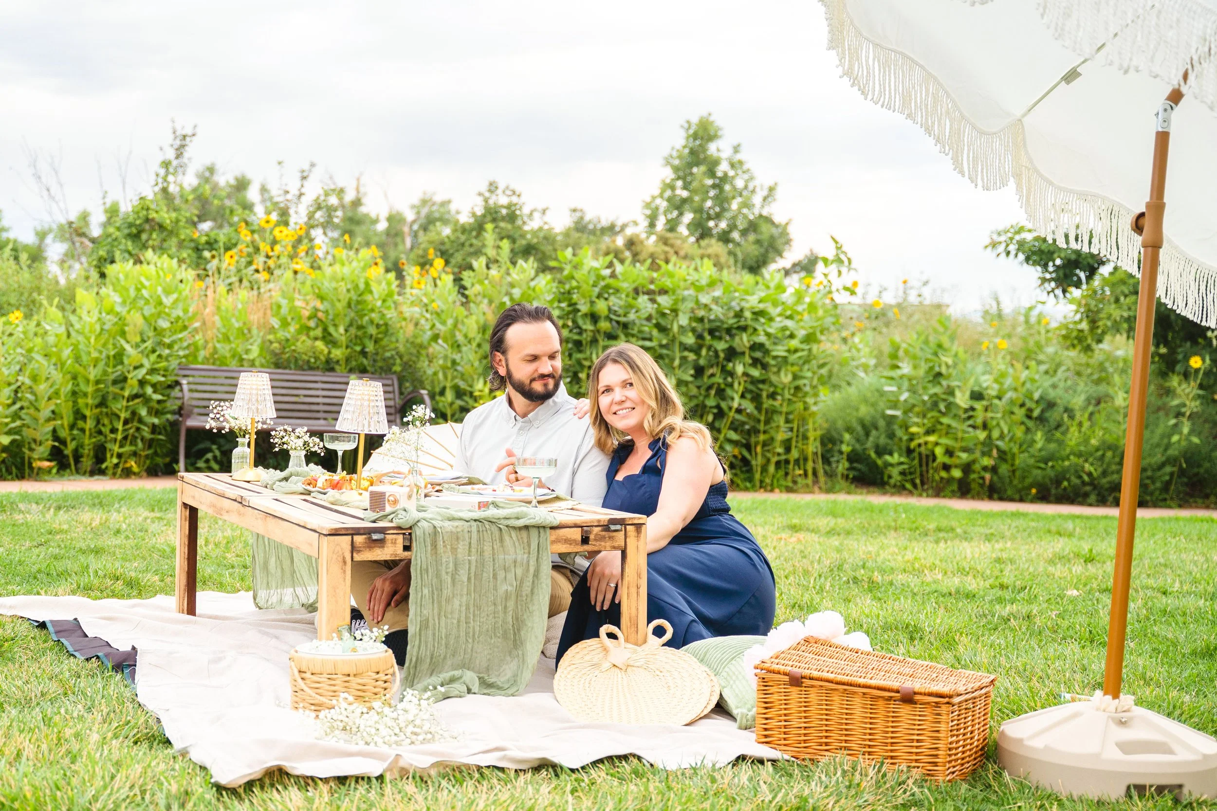 A couple at a picnic table outdoors, under a large white umbrella, with greenery and yellow flowers in the background.
