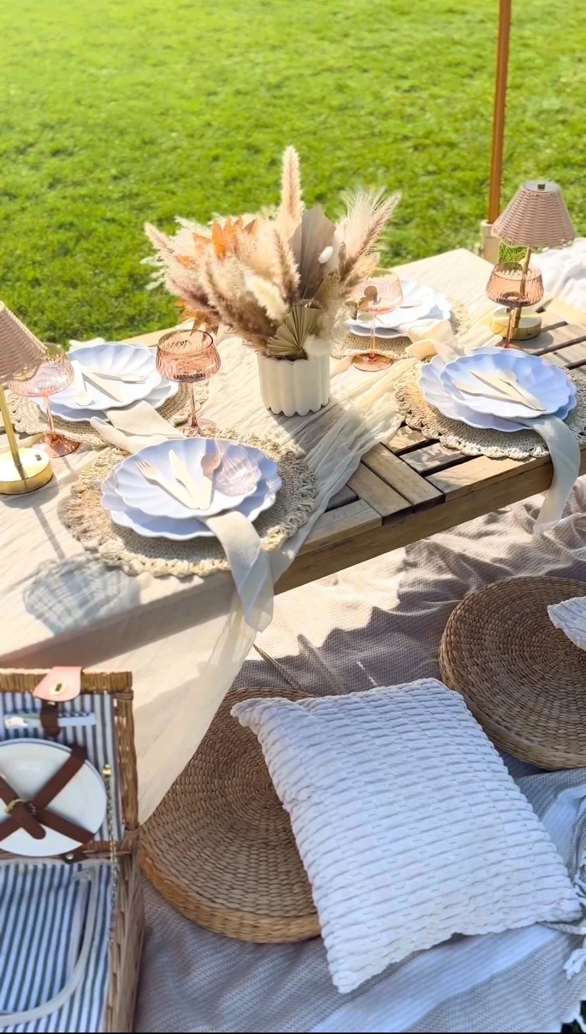 Outdoor luxury picnic table setup with white tableware, a vase of dried pampas grass, pink lamps, wicker chairs, and cushions, on a grassy lawn in City Park, Denver.