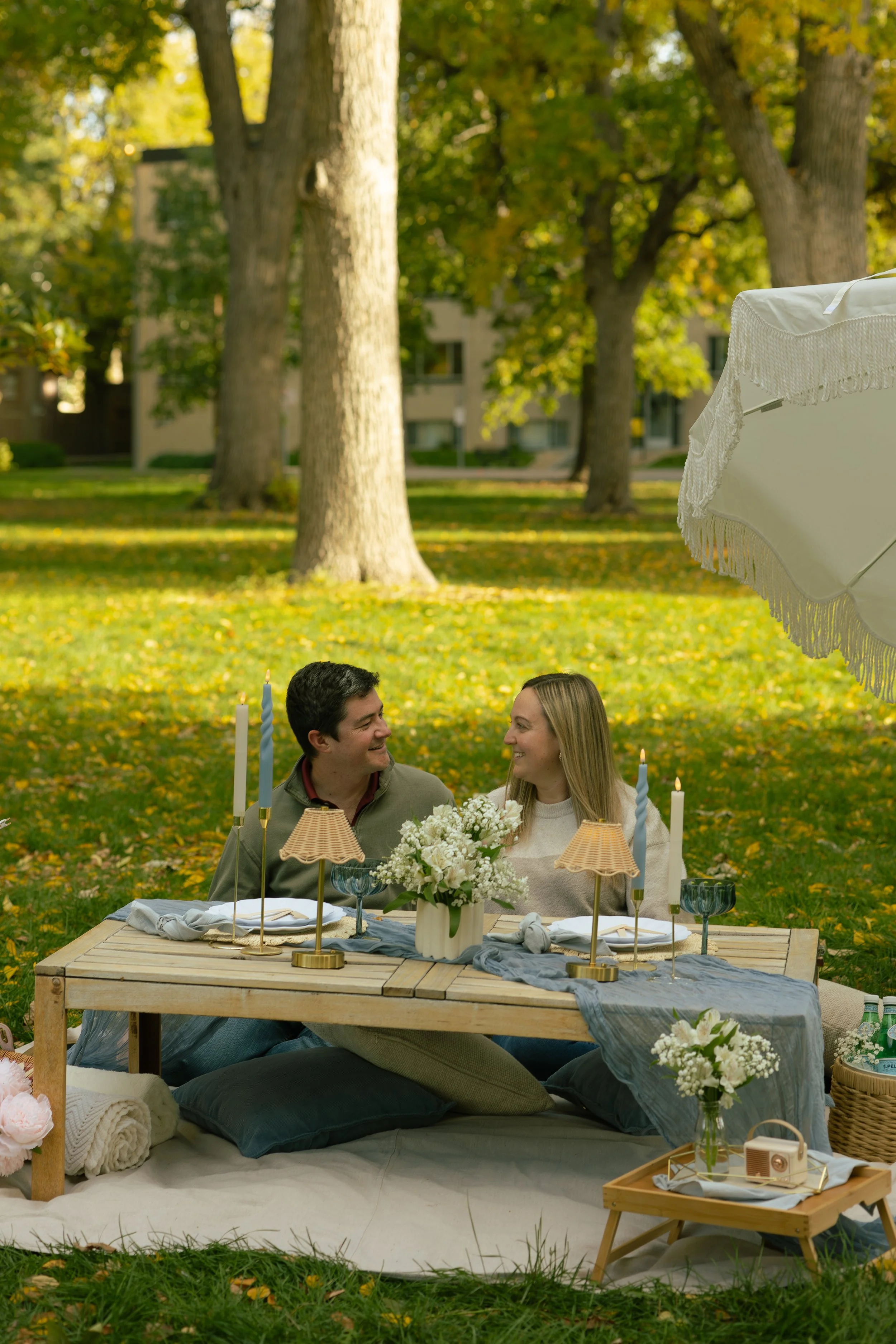 A couple sitting at a table outdoors during daytime, surrounded by trees with autumn foliage, celebrating a special occasion with floral arrangements, candles, and table settings.