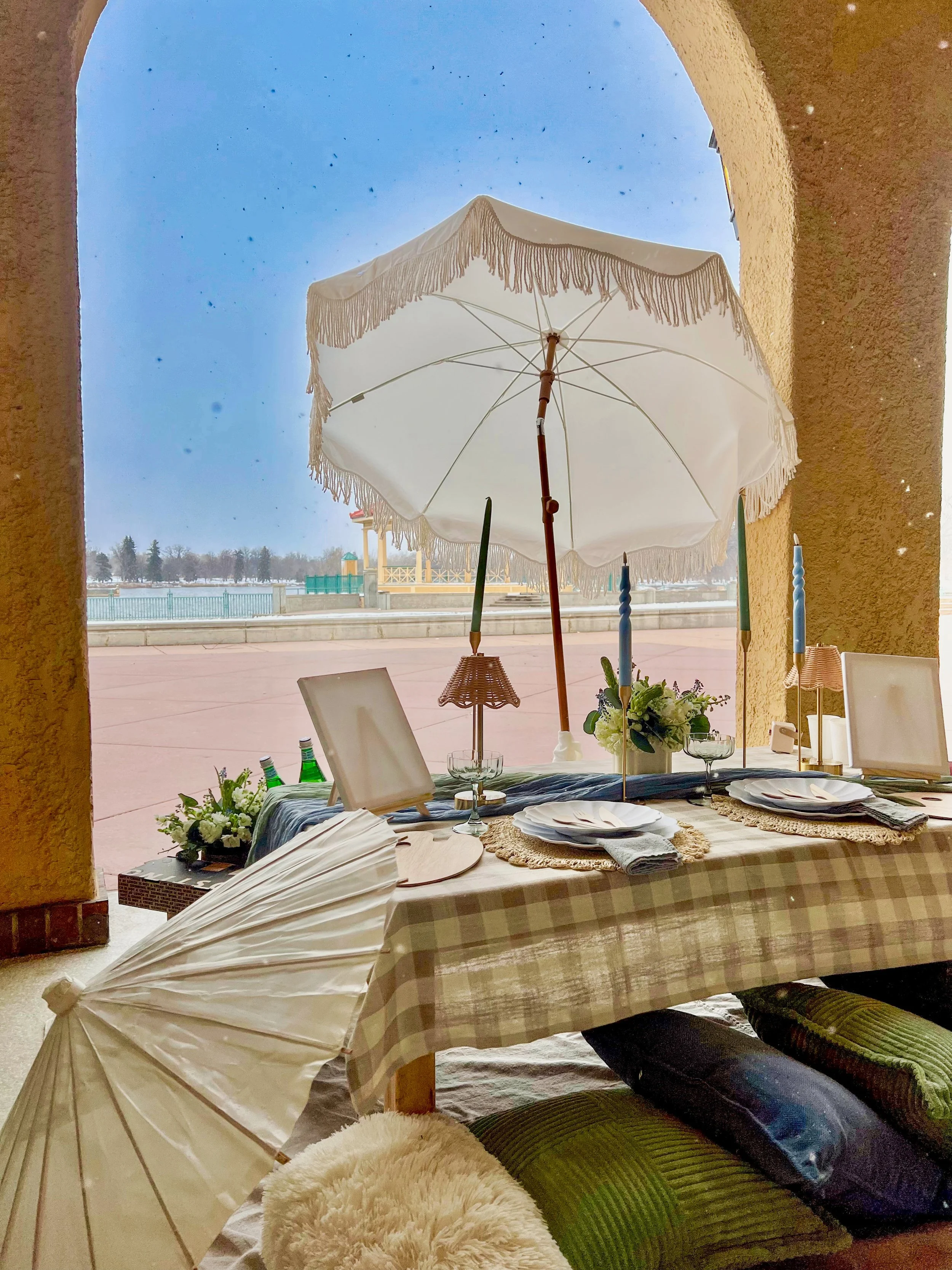 Indoor scene with a table set for a meal, decorated with flowers, plates, napkins, and candles. A large white umbrella is propped open outside on a patio, with a view of a park or waterfront area in the background under a clear blue sky.