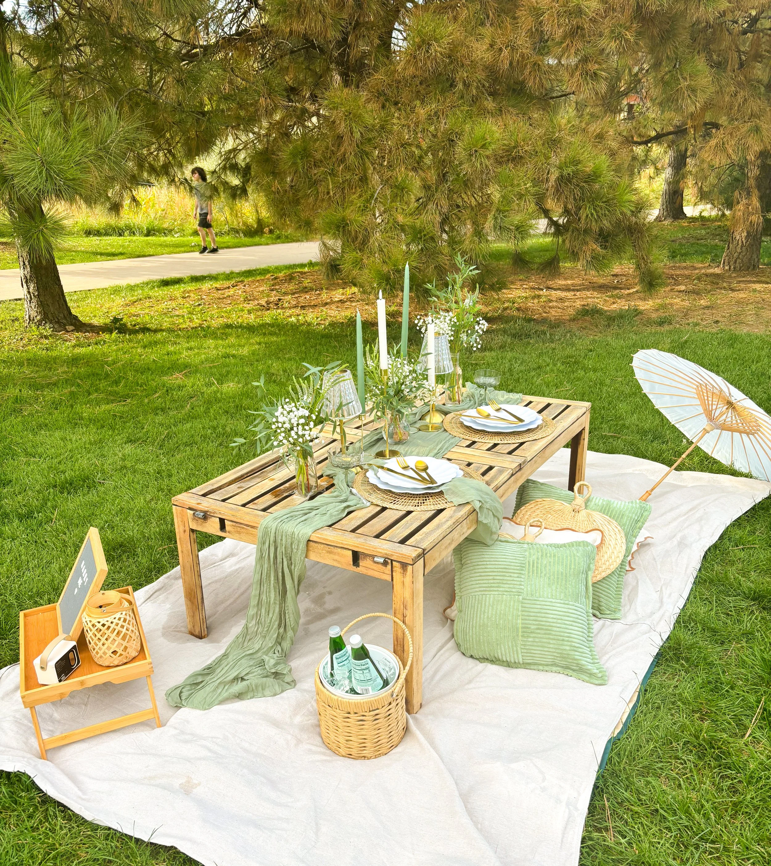 Outdoor table set for a picnic with white plates, gold utensils, glass cups, green cloth napkins, candles, and small flower arrangements on a wooden table. A green cushion and a large green and white striped pillow are on a blanket on the grass. A wi