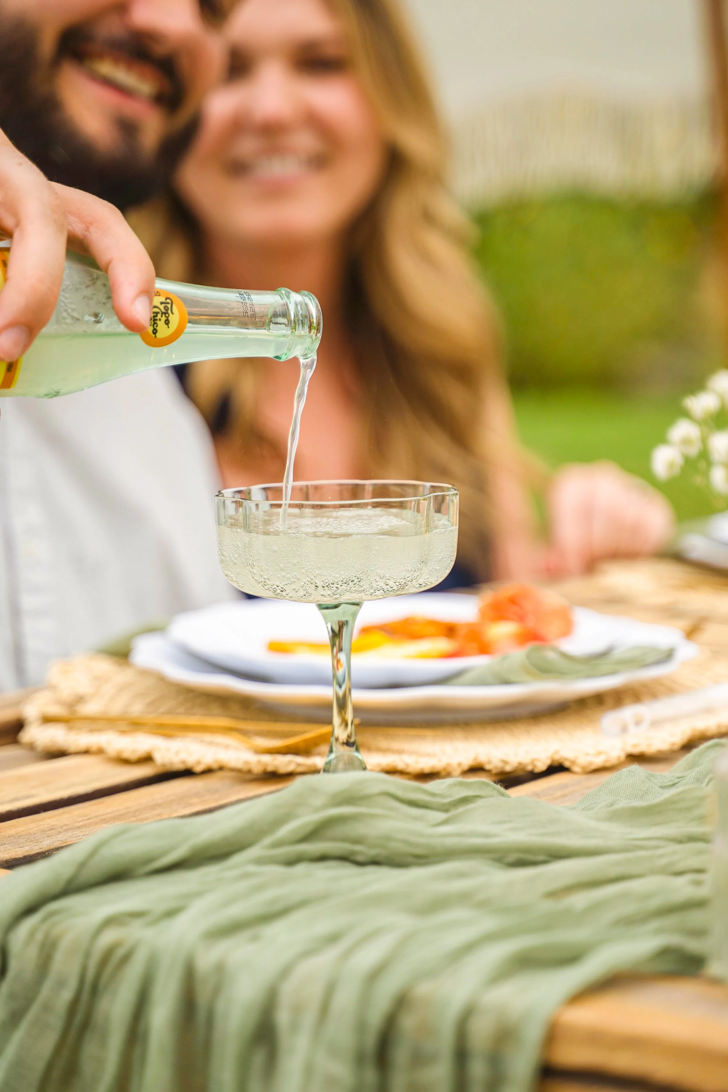 A woman pouring a clear drink from a glass bottle into a cocktail glass at an outdoor meal setting, with a smiling man and woman in the background.