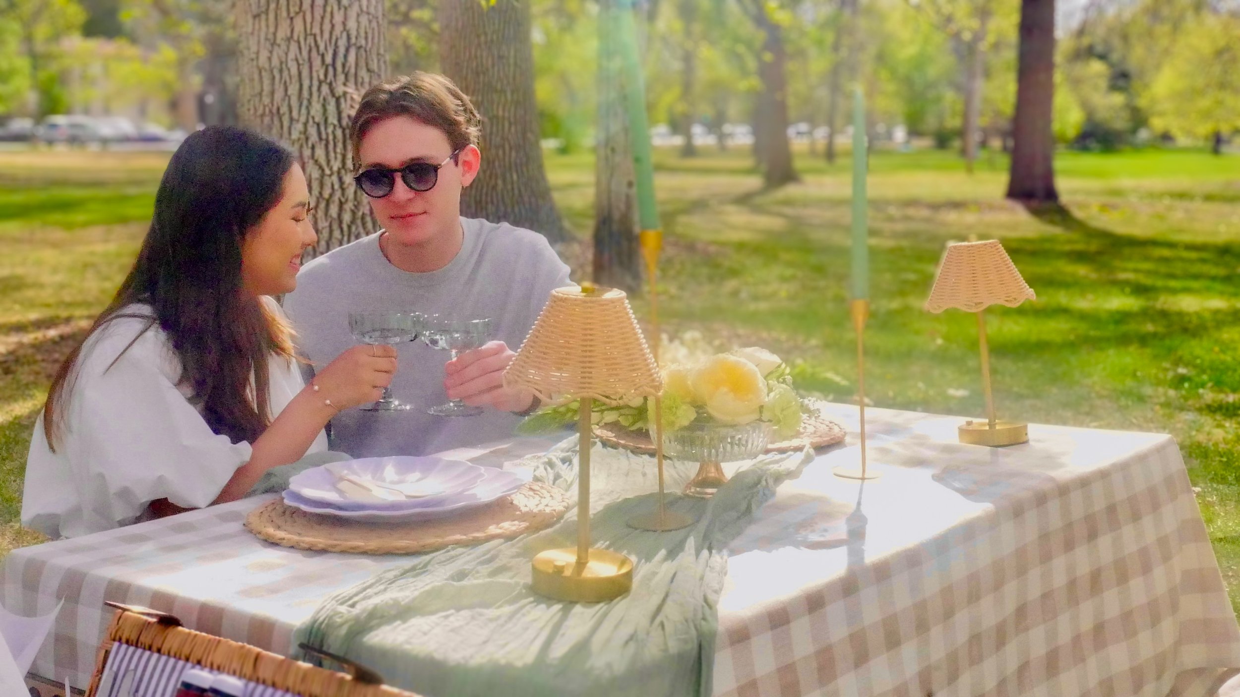 Two people enjoying an outdoor meal at a luxury picnic table in Denver City Park with green grass and trees. One person is wearing sunglasses, and they are toasting with drinks.