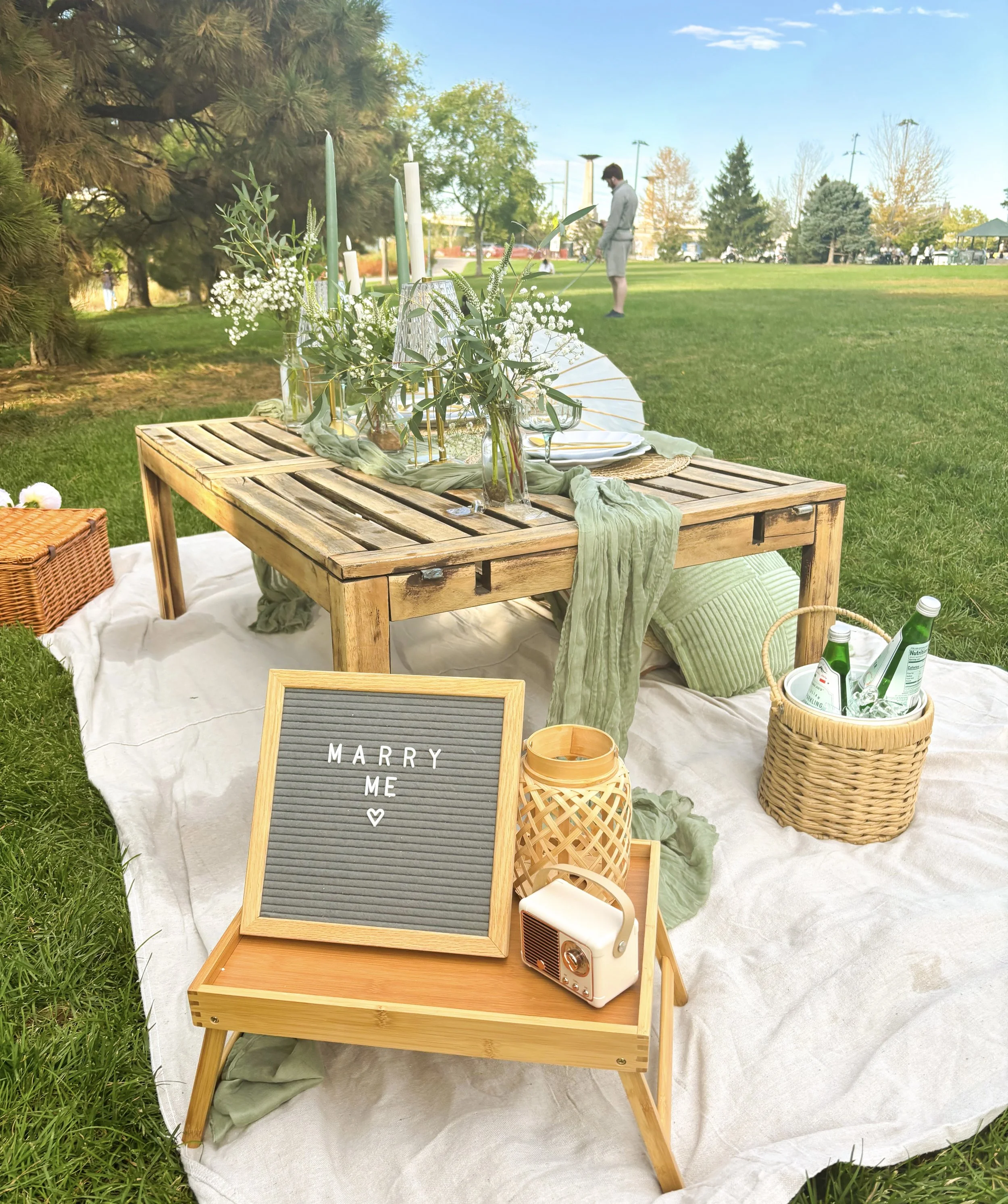 Outdoor luxury picnic proposal setup with a wooden table decorated with candles and flowers, placed on a white cloth, with a chalkboard sign reading 'Marry Me' and a small speaker nearby, on lush green grass in a park.