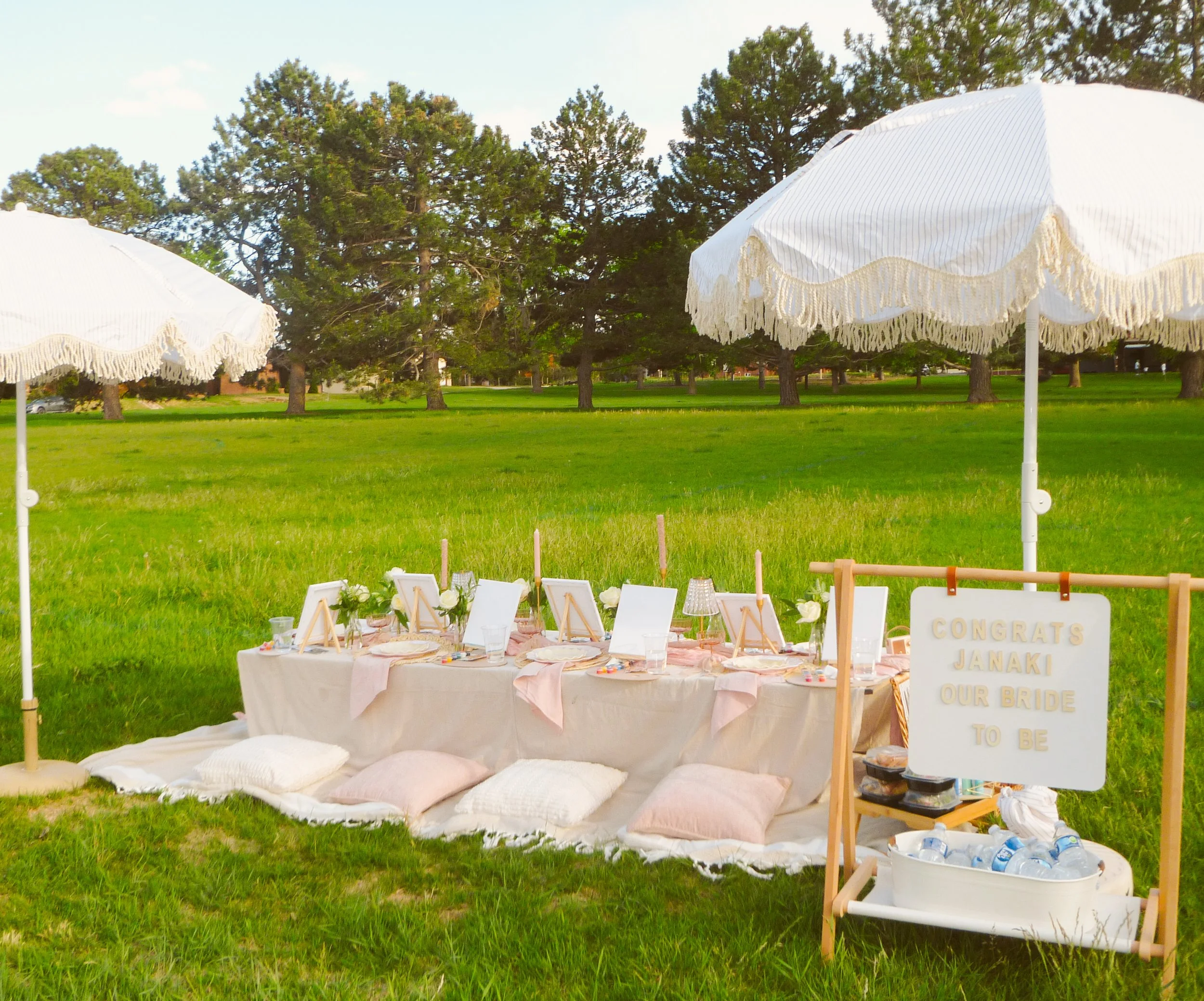 Outdoor bachelorette picnic setup with white umbrellas, a decorated table with pillows, framed photos, and a congratulatory sign for Janaki in Wash Park, Denver.