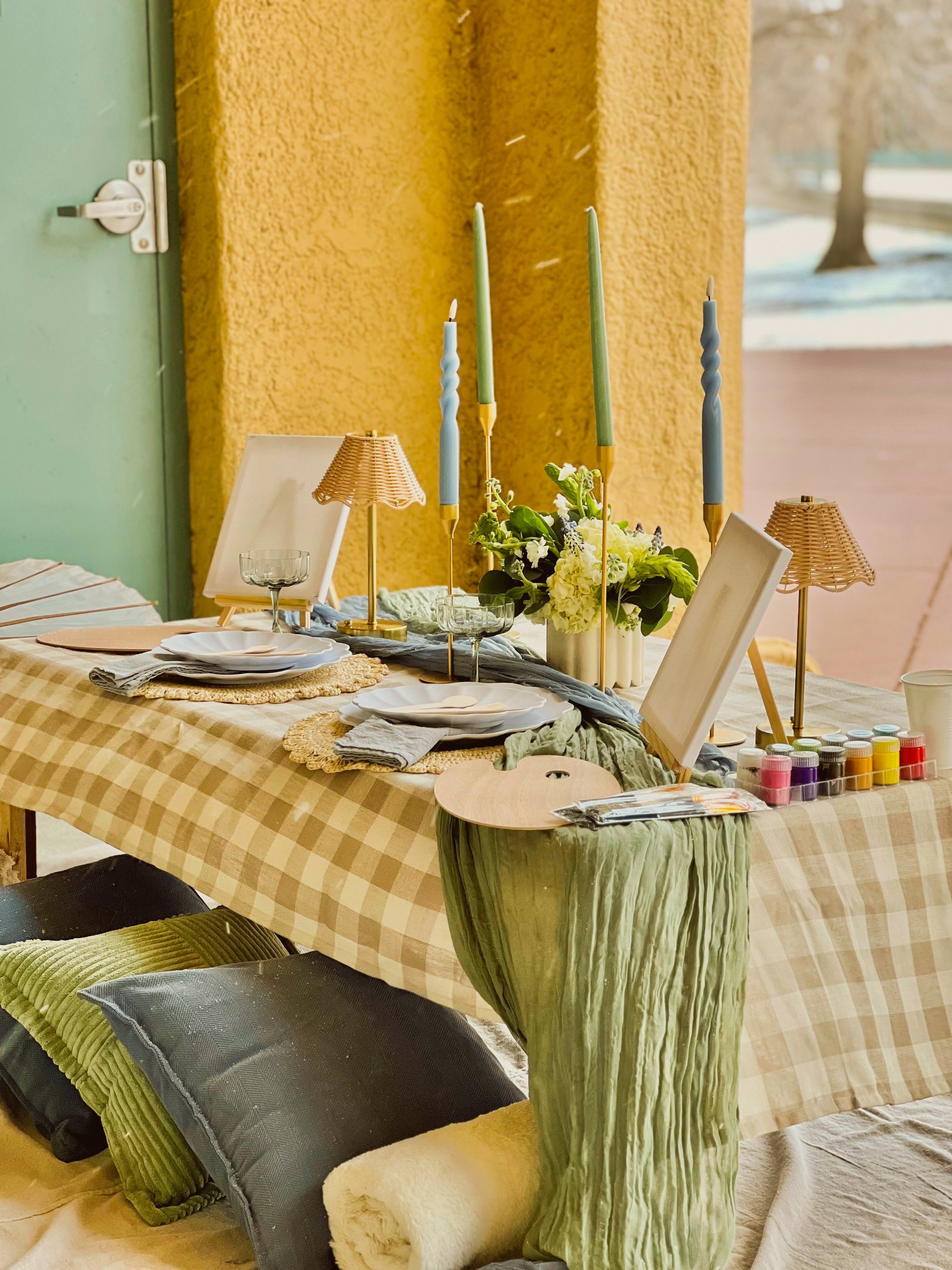 A decorated table with candles, flowers, plates, bowls, and lamps on a checkered tablecloth. There are cushions on the floor in front of the table and a large green vase in the foreground. The background shows a yellow textured wall.