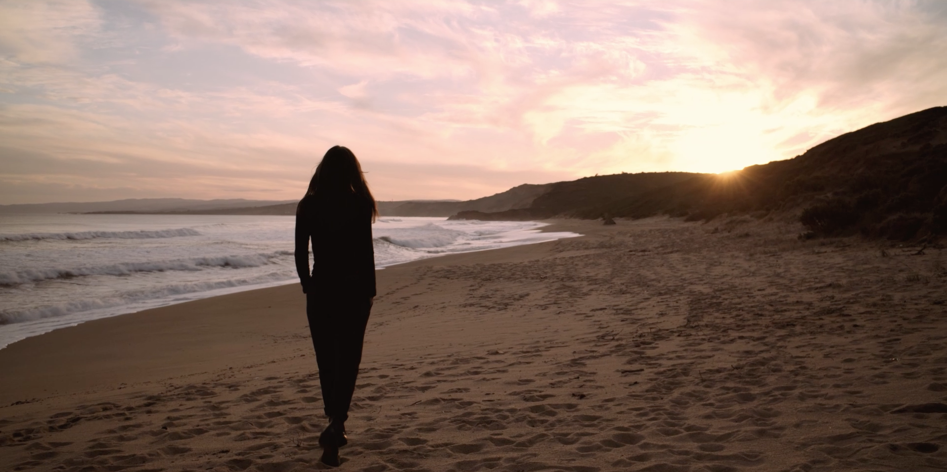 A person walking alone on a sandy beach during sunset, with waves crashing and sand dues in the background.