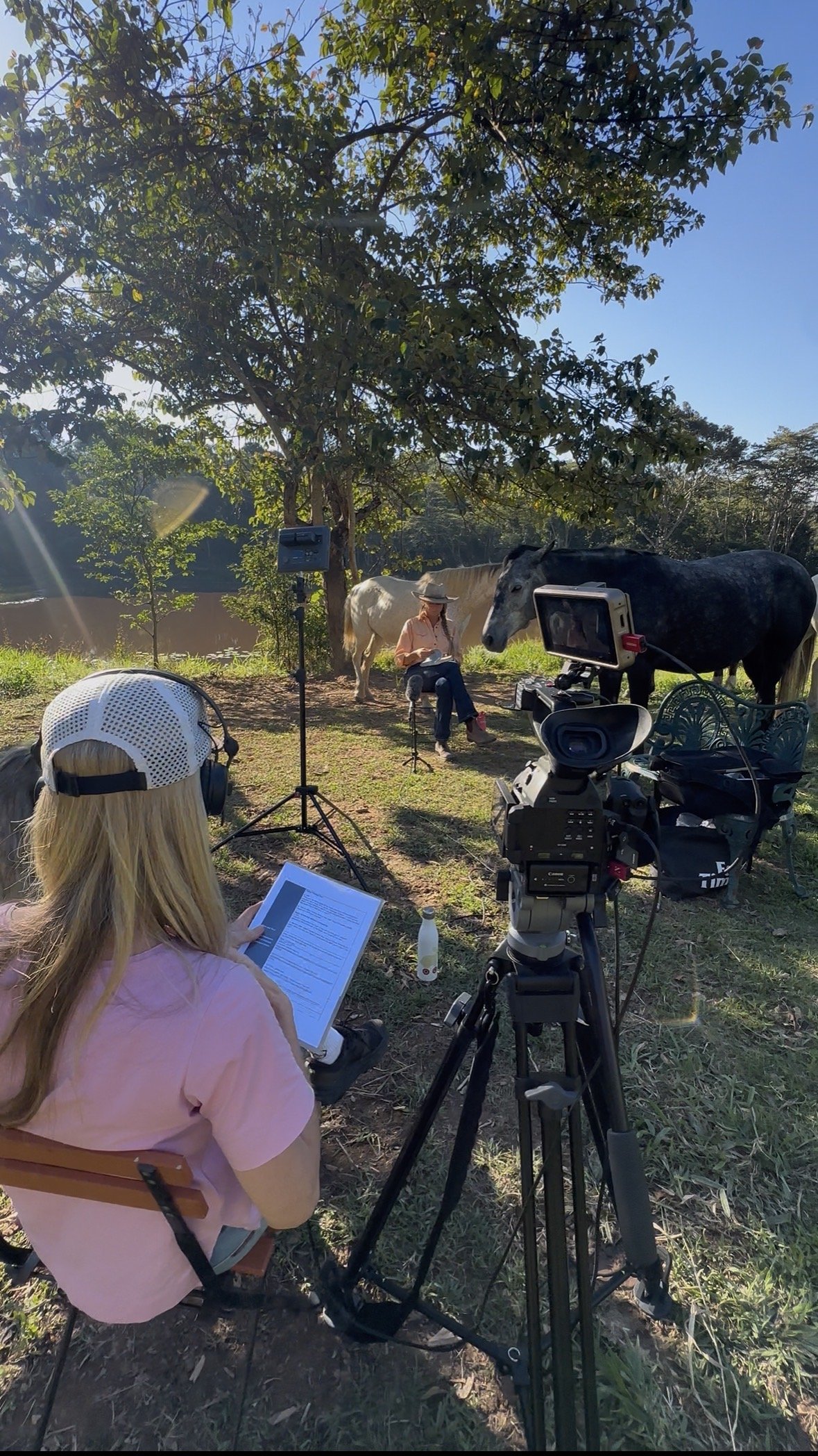 A woman sitting outdoors with a notebook, recording a scene involving a horse and a person under a tree, with a camera on a tripod recording the scene.