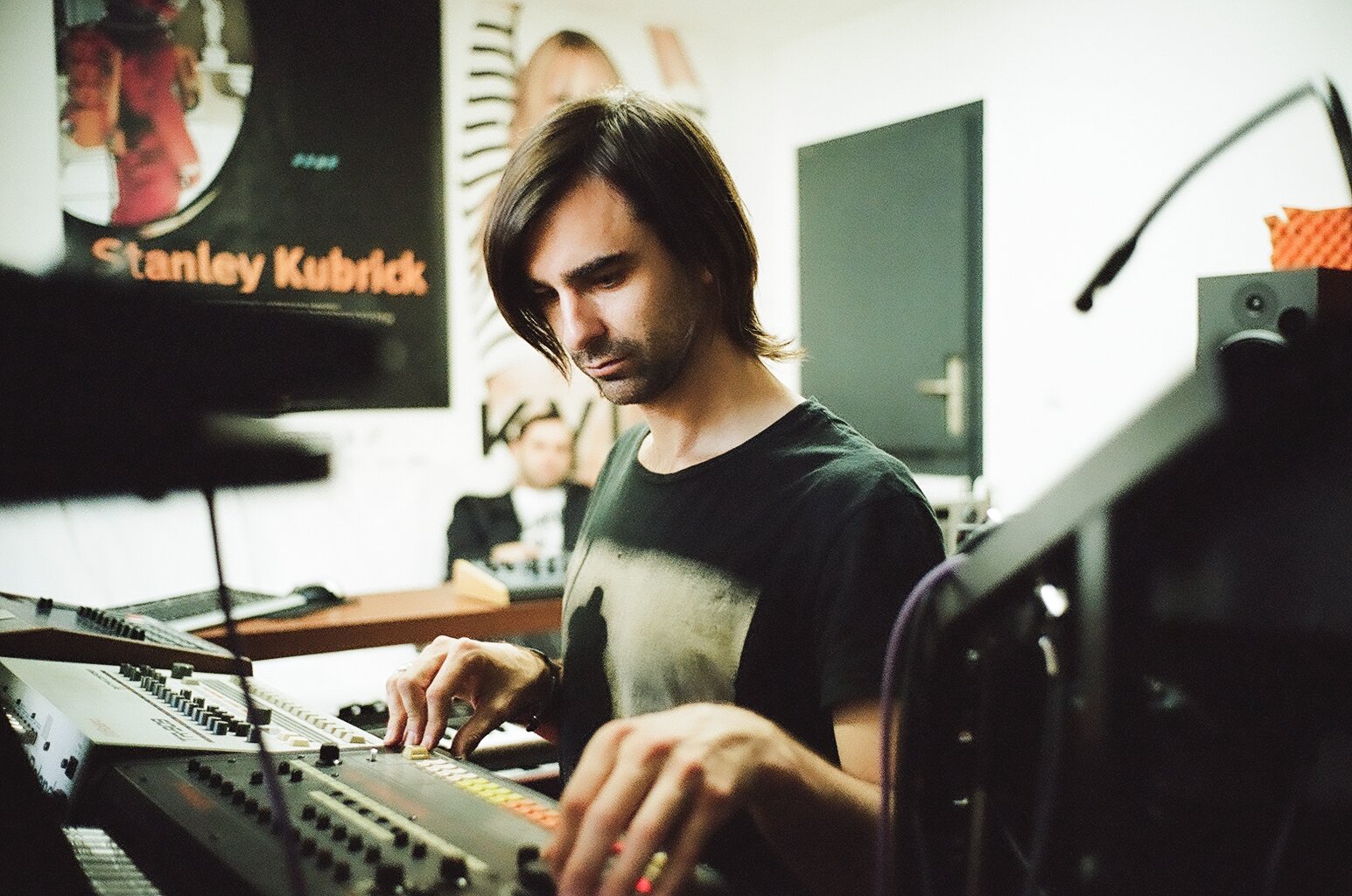 A young man with long dark hair and a black t-shirt is playing a synthesizer keyboard in a music studio.