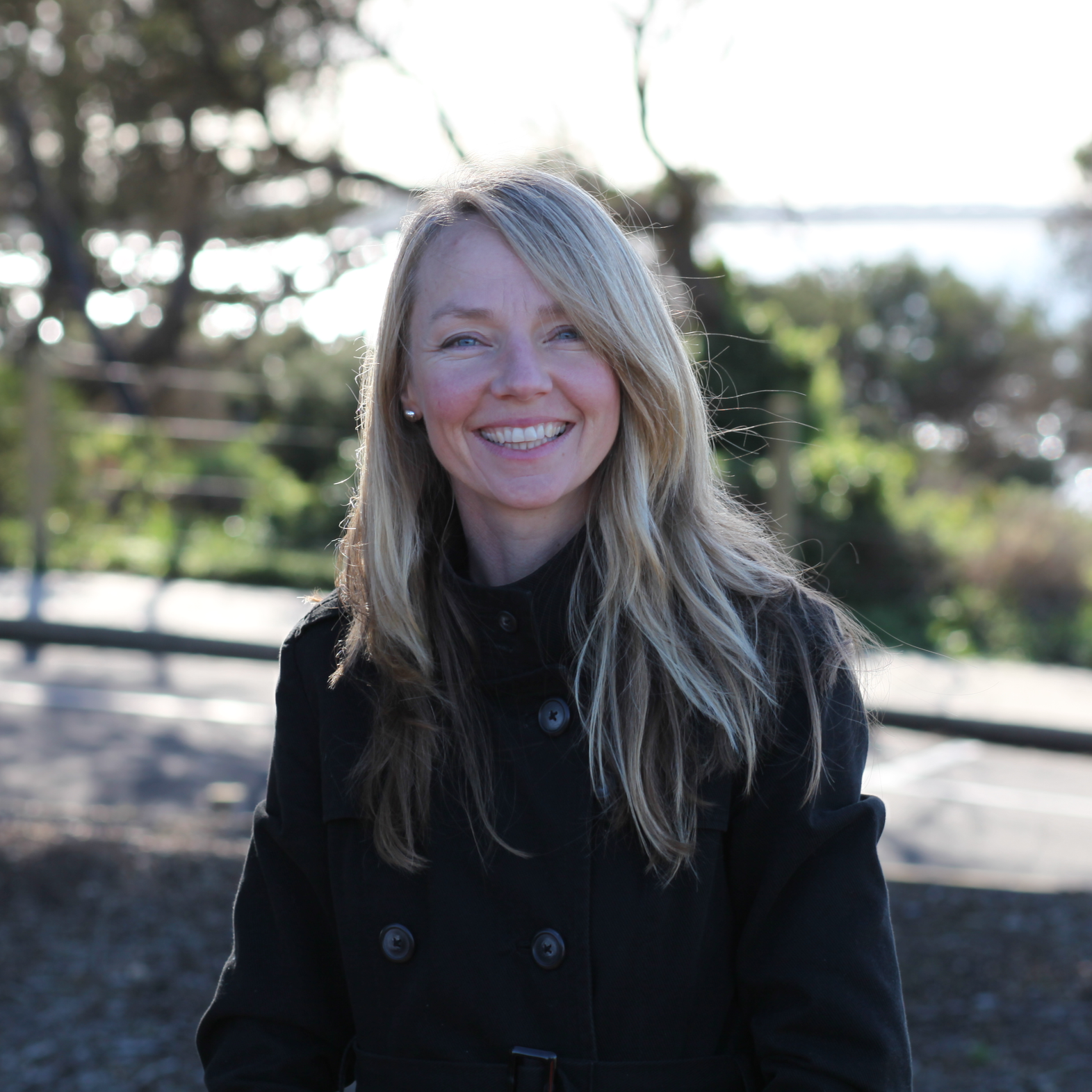 A woman with long blonde hair, smiling, wearing a black jacket, outdoors with trees and water in the background.