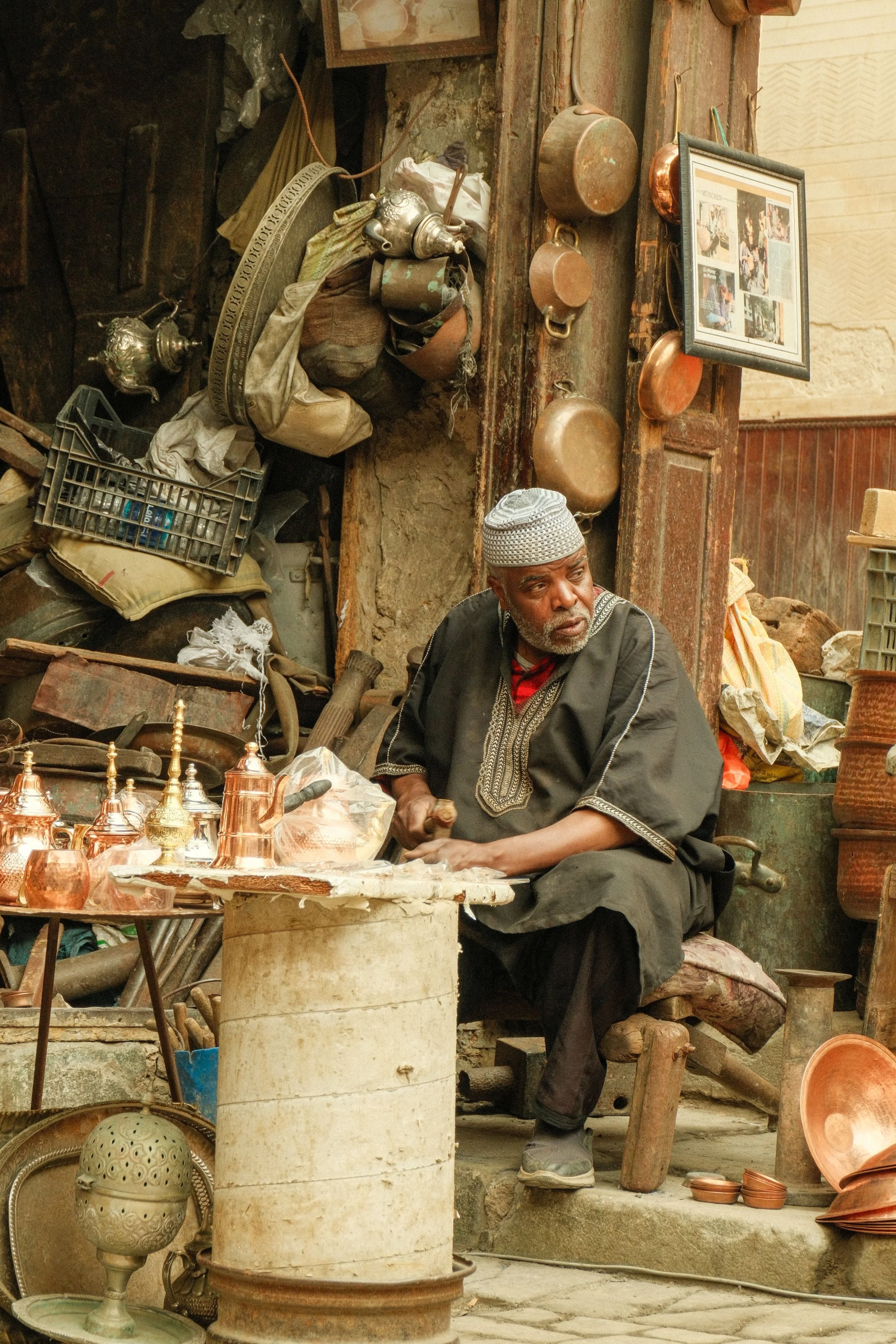 A man wearing traditional attire and a cap, sitting at a market stall with various metallic and ceramic items, including teapots, bowls, and decorative pieces, in a rustic setting with hanging pots, framed photographs, and cluttered surroundings.