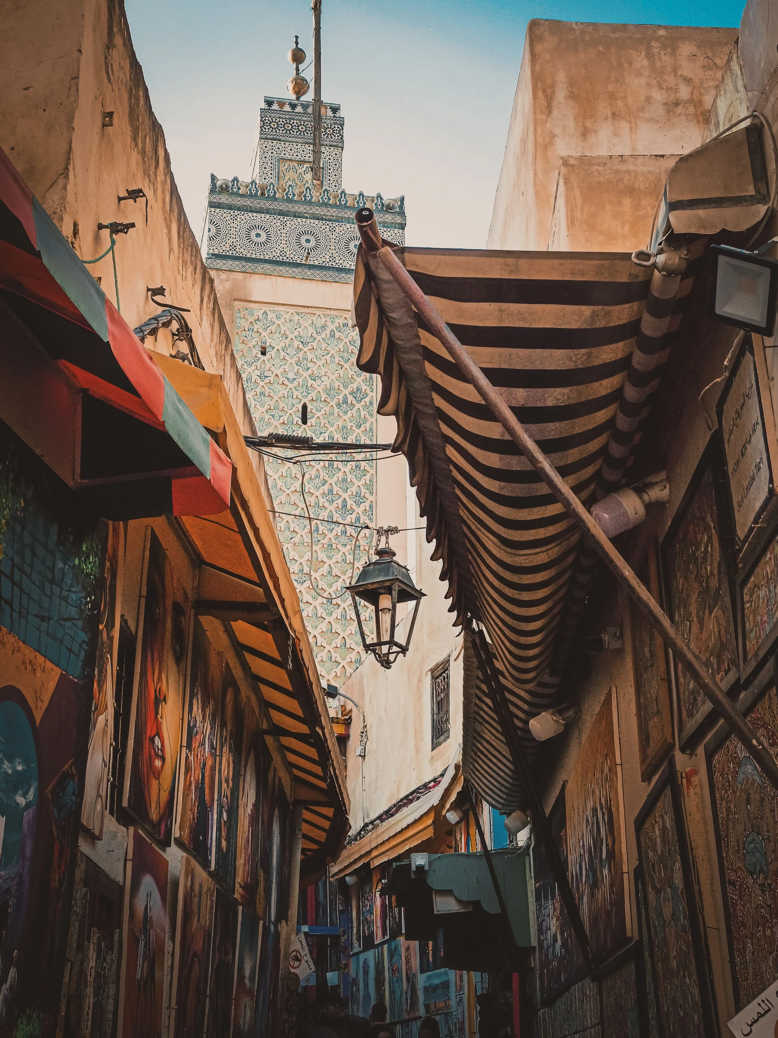 Tall minaret with Islamic geometric tiles at the top of a mosque, seen from a narrow street with walls covered in street art and paintings.