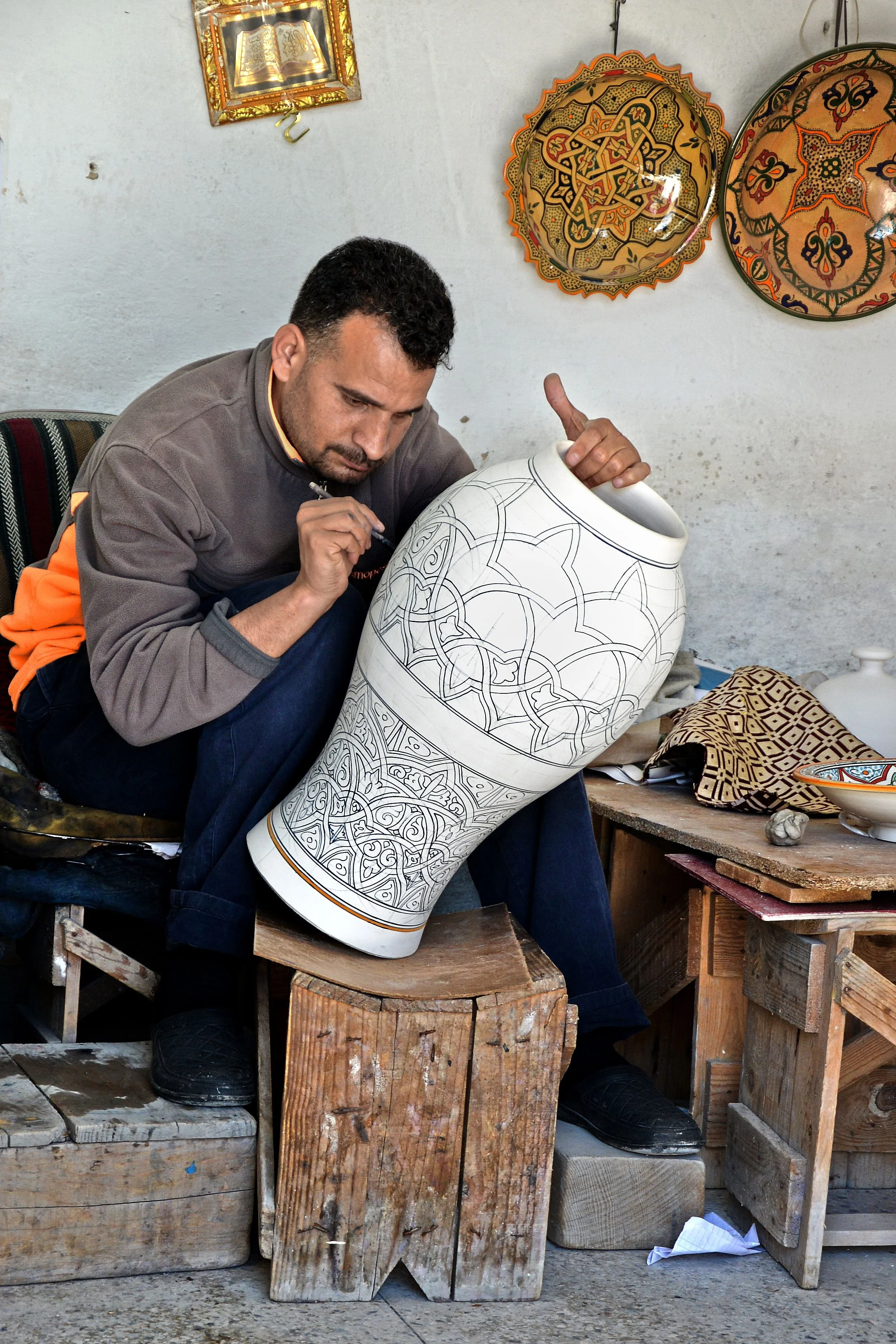 A man sitting outdoors on a wooden stool, intricately painting a large, decorated ceramic vase with detailed patterns. The setting includes wooden blocks and a table with various art supplies and decorative items, with colorful, patterned plates hanging on the wall above.