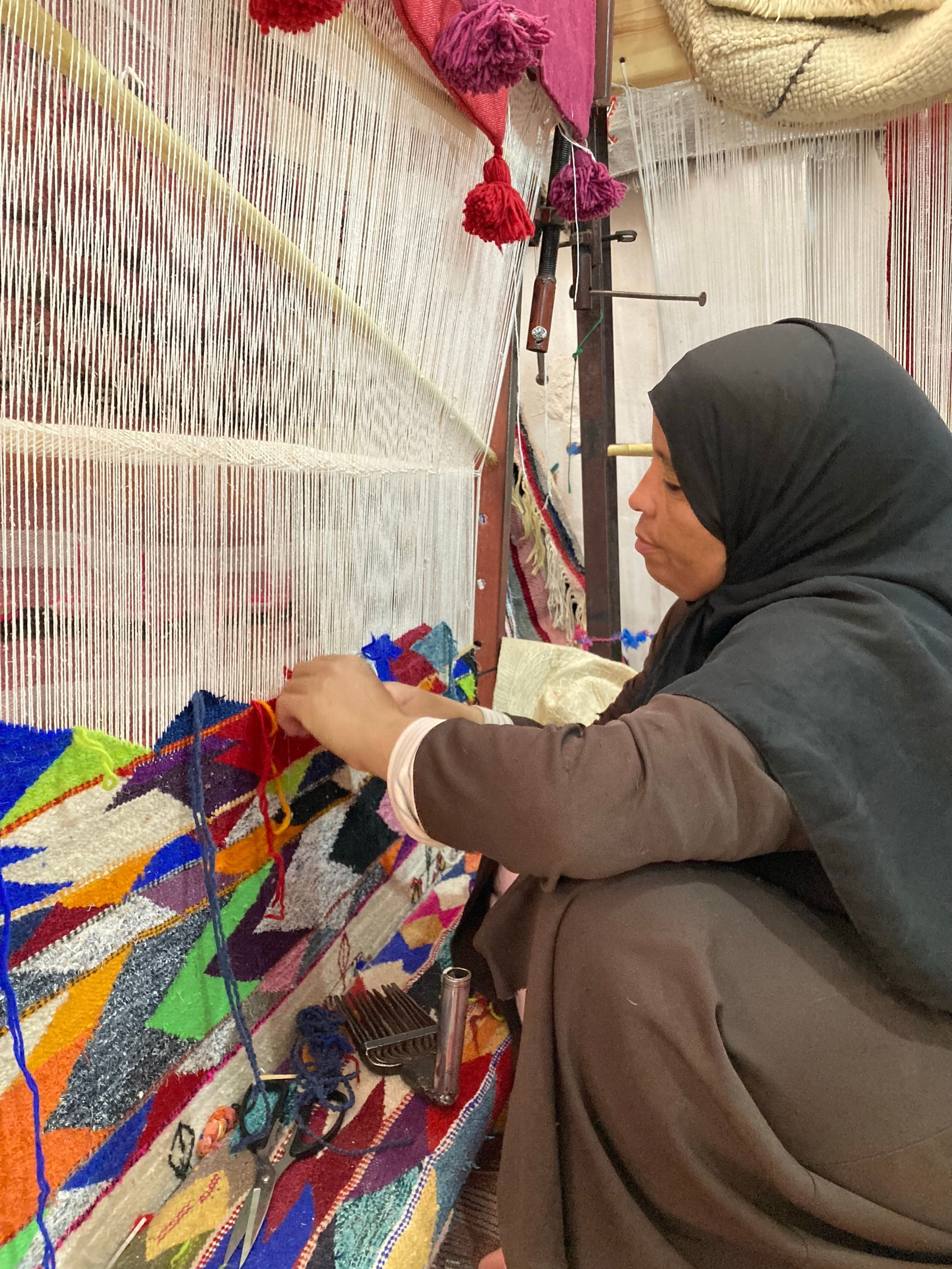A woman with a black headscarf working on a colorful, woven tapestry, using a weaving loom in a workshop.
