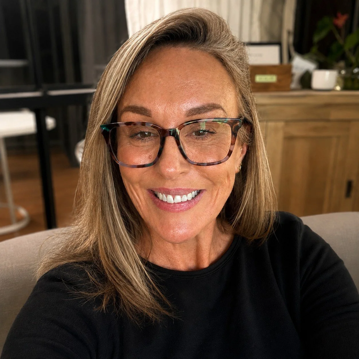 A smiling woman with blonde hair and glasses taking a selfie indoors.