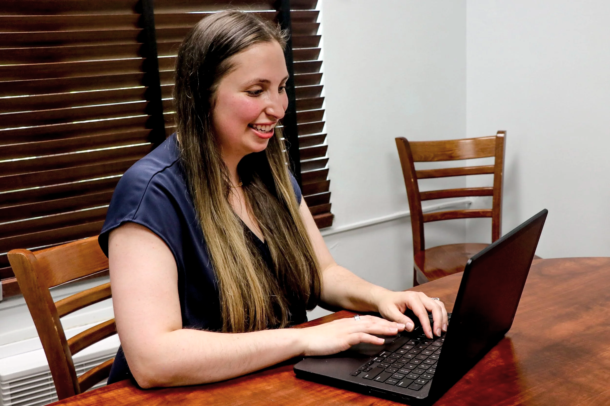 A young woman with long brown hair smiling and working on a black laptop at a wooden table, with wooden chairs and a window with closed blinds in the background.