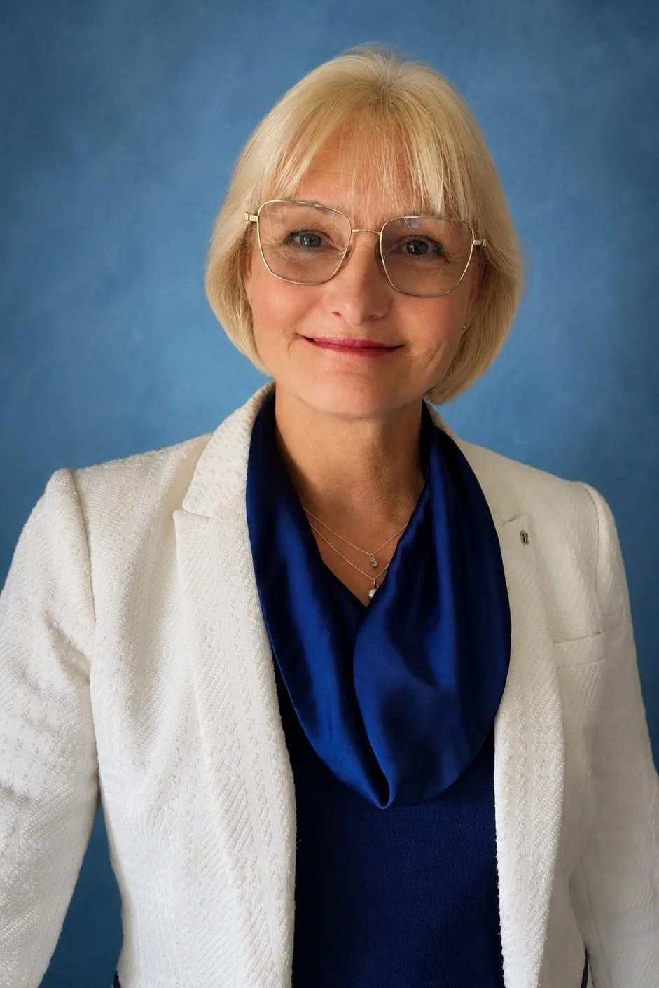 A woman with short blonde hair, glasses, and light skin, smiling in front of a blue background. She is wearing a white jacket, a dark blue blouse with a silk scarf, and layered necklaces.
