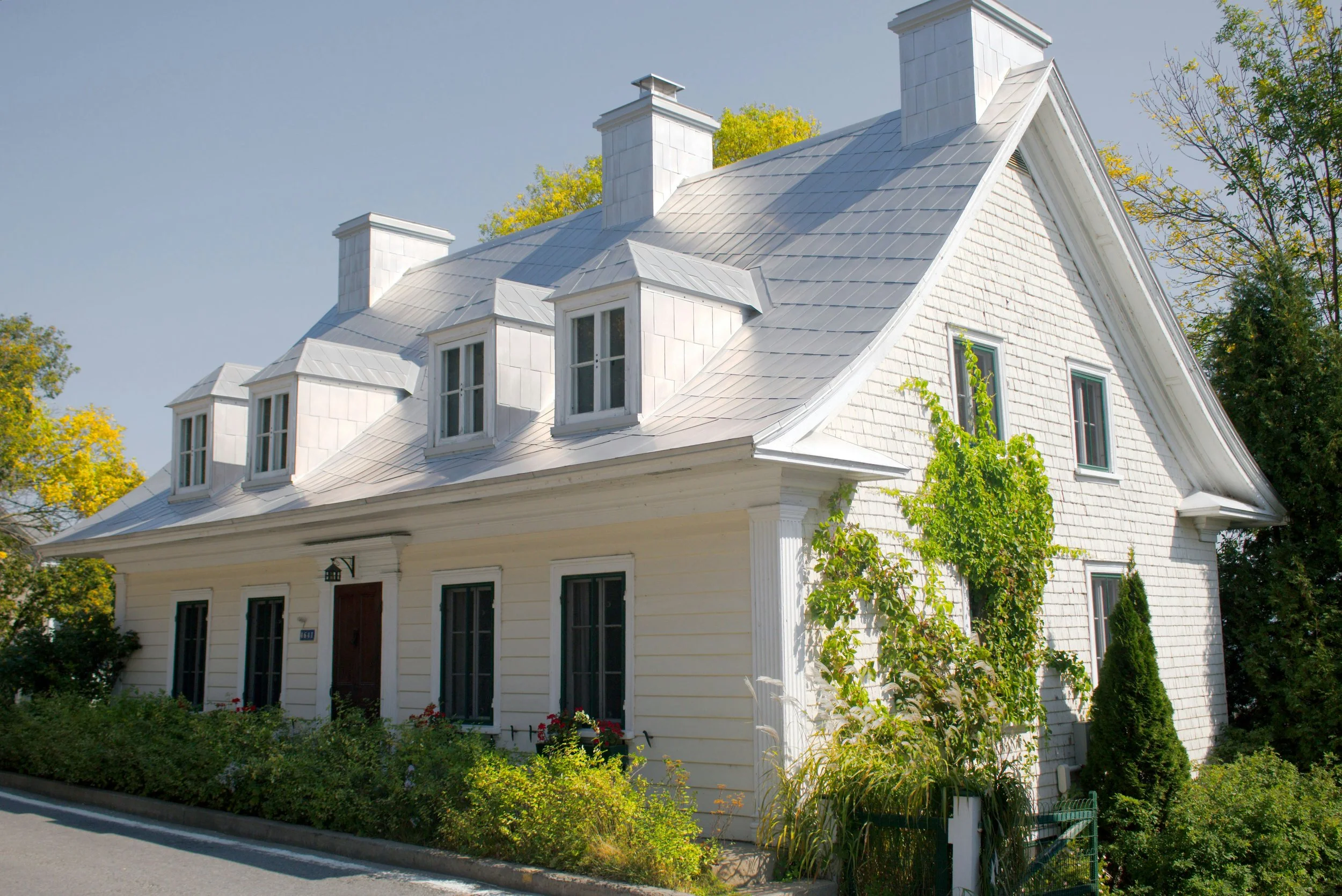 A white house with a steep roof, multiple dormer windows, and chimneys, surrounded by greenery and trees under a clear sky.