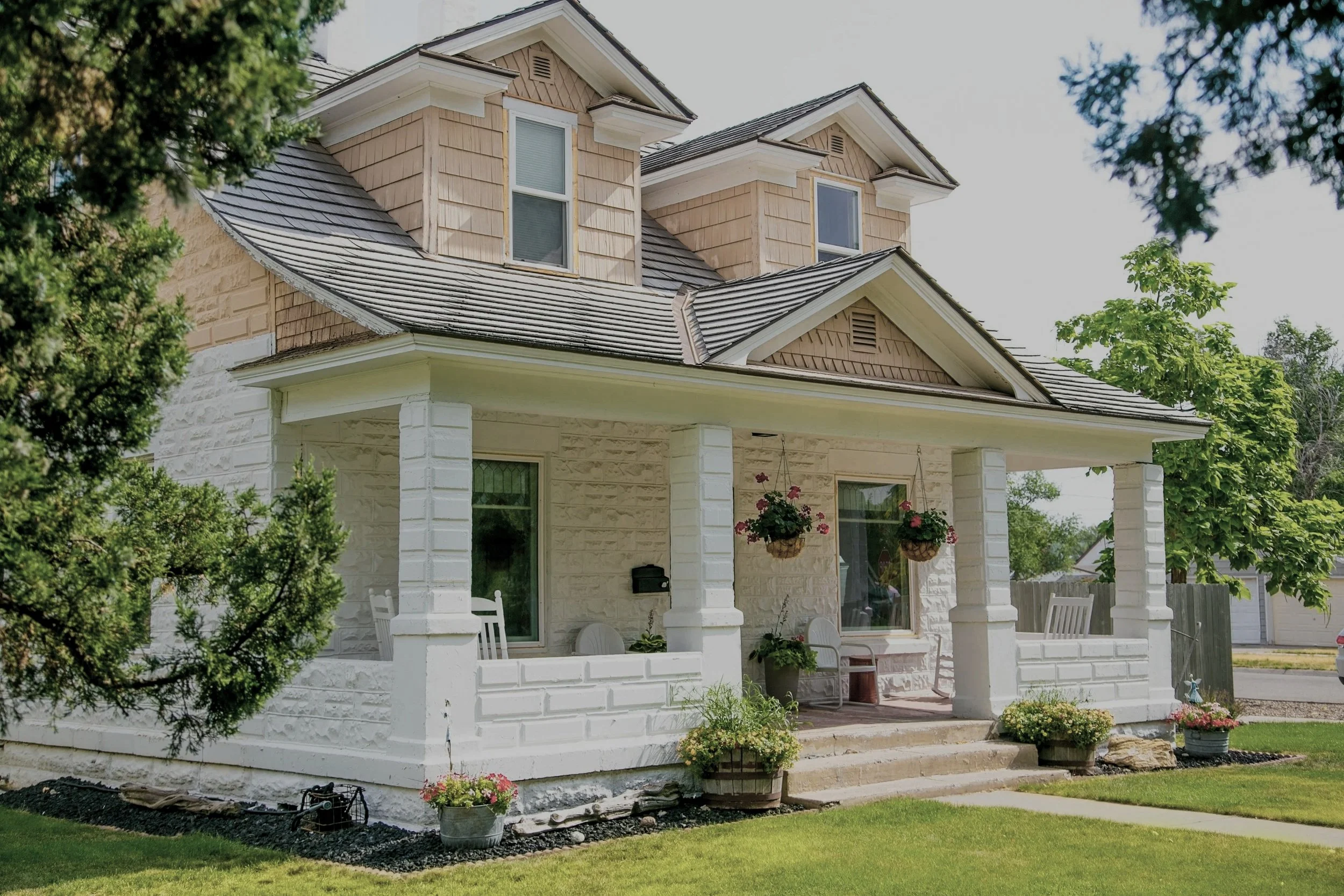 A two-story house with beige shingles, white brick exterior, and a covered porch. The house has two upper windows and hanging flower baskets, surrounded by green trees and a lawn.