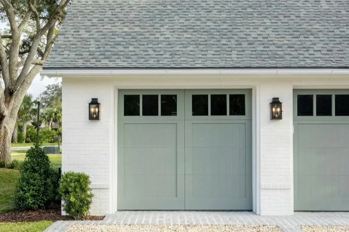 Exterior of a modern garage with gray double doors, white brick walls, and black wall-mounted lantern-style lights on either side of the doors. There are green bushes and trees in the background.