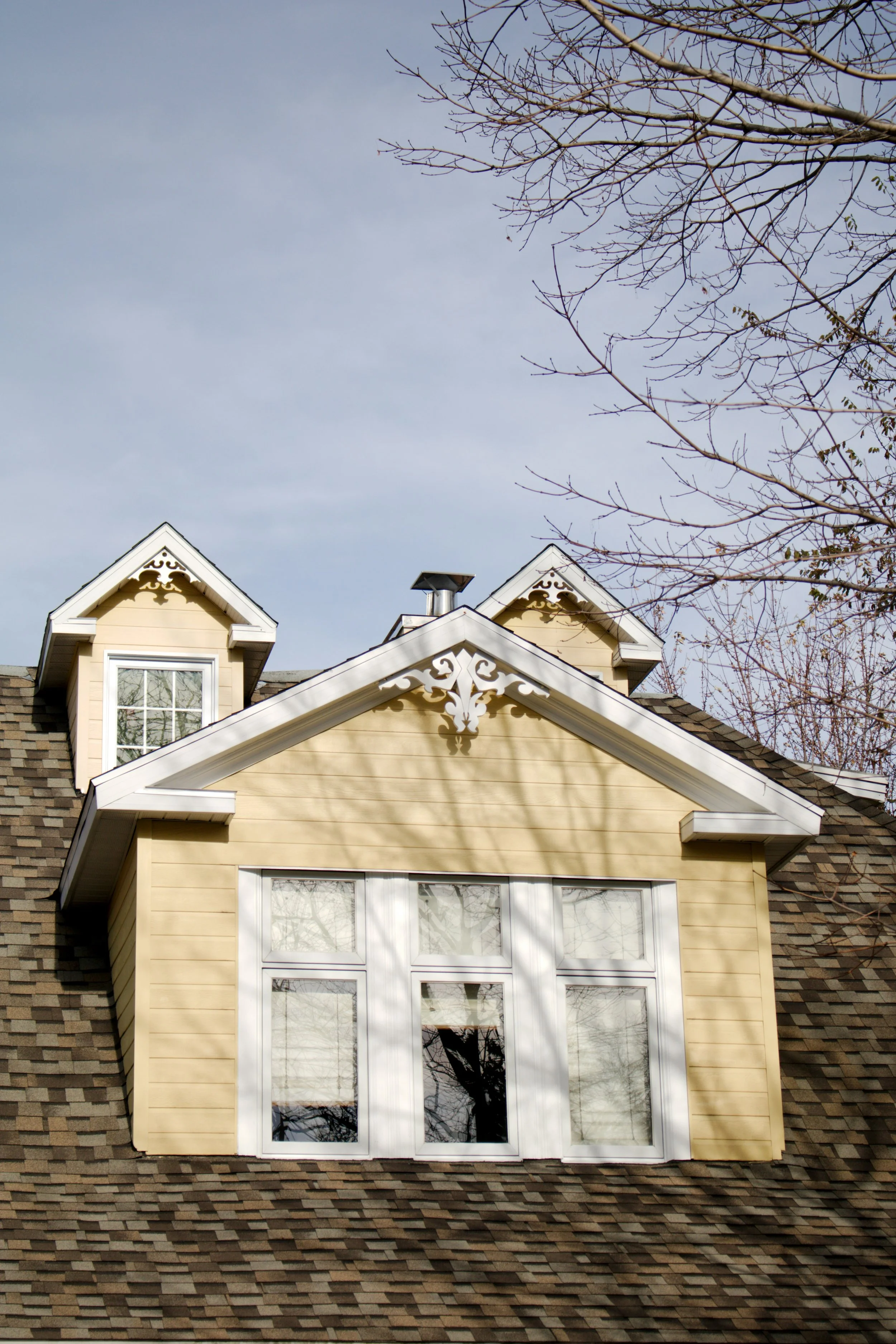 Yellow house with white decorative trim and a large window on the upper floor, with a leafless tree in the foreground and part of the roof visible.