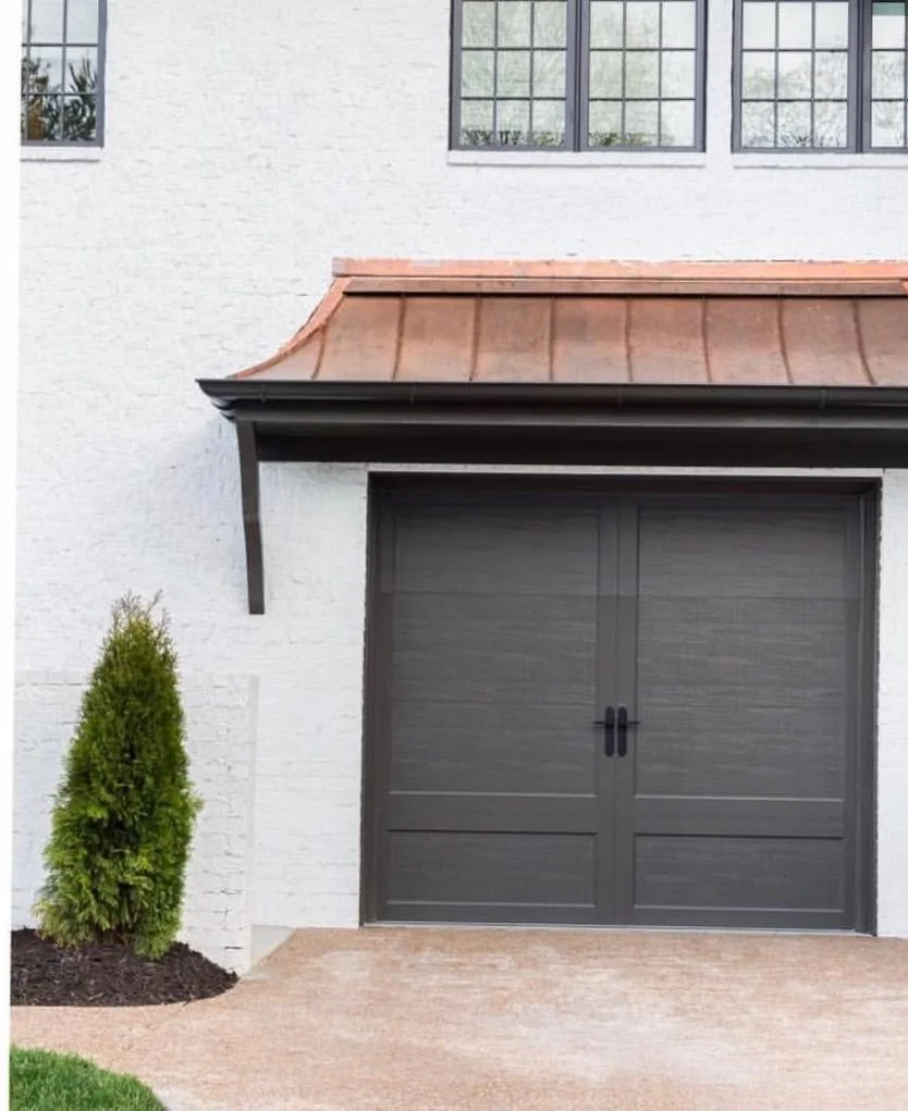 A modern house with a white brick exterior, black garage doors, small front yard with a green bush, and a copper-colored roof above the garage.