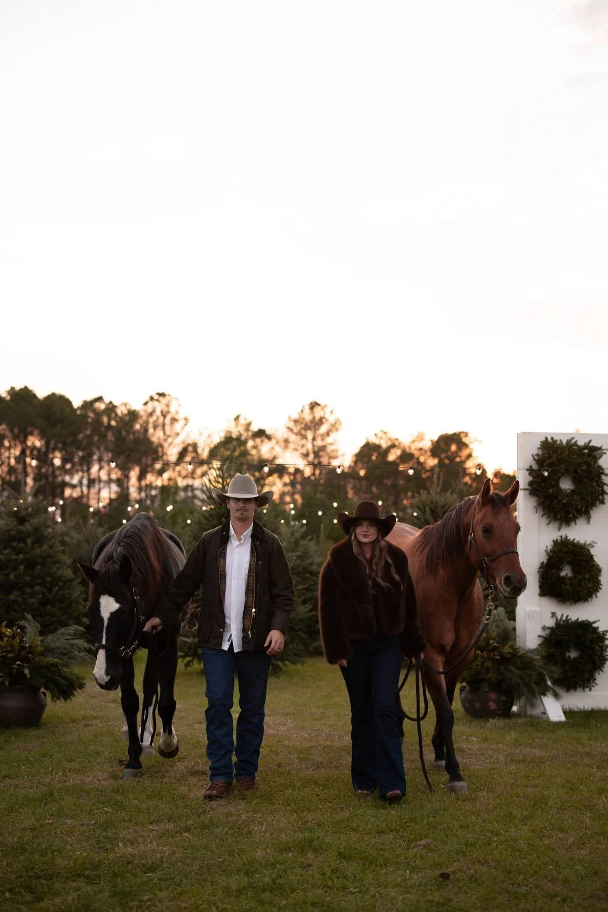 A man and woman walking with horses during a festive outdoor event at sunset, surrounded by Christmas greenery and decorations.