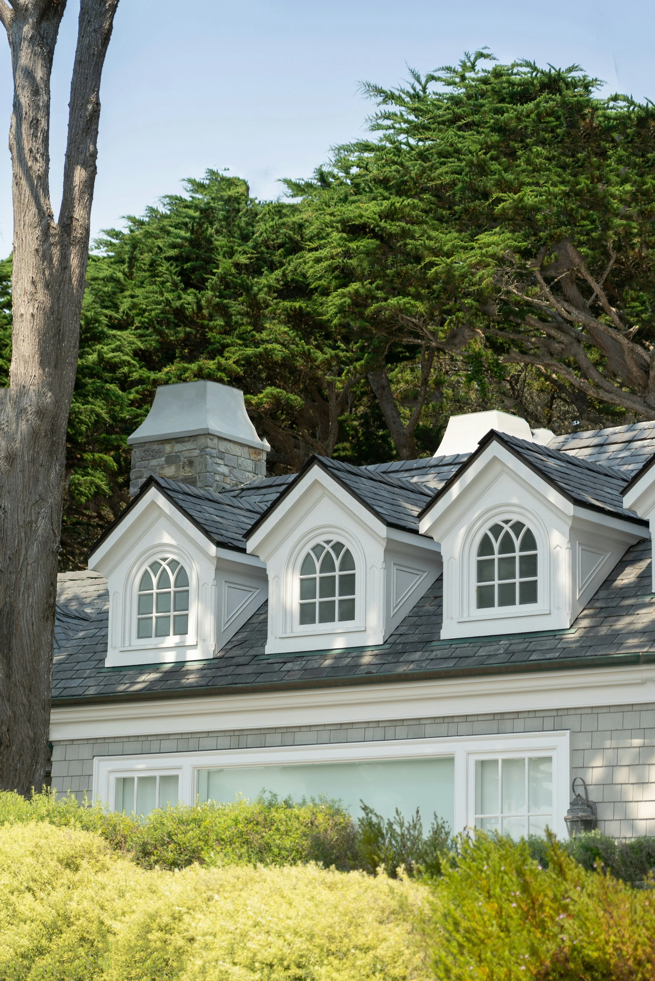 The image shows a house with a Tudor-style roof featuring multiple dormer windows, a stone chimney, and gray shingle siding, surrounded by lush greenery and tall trees in the background.