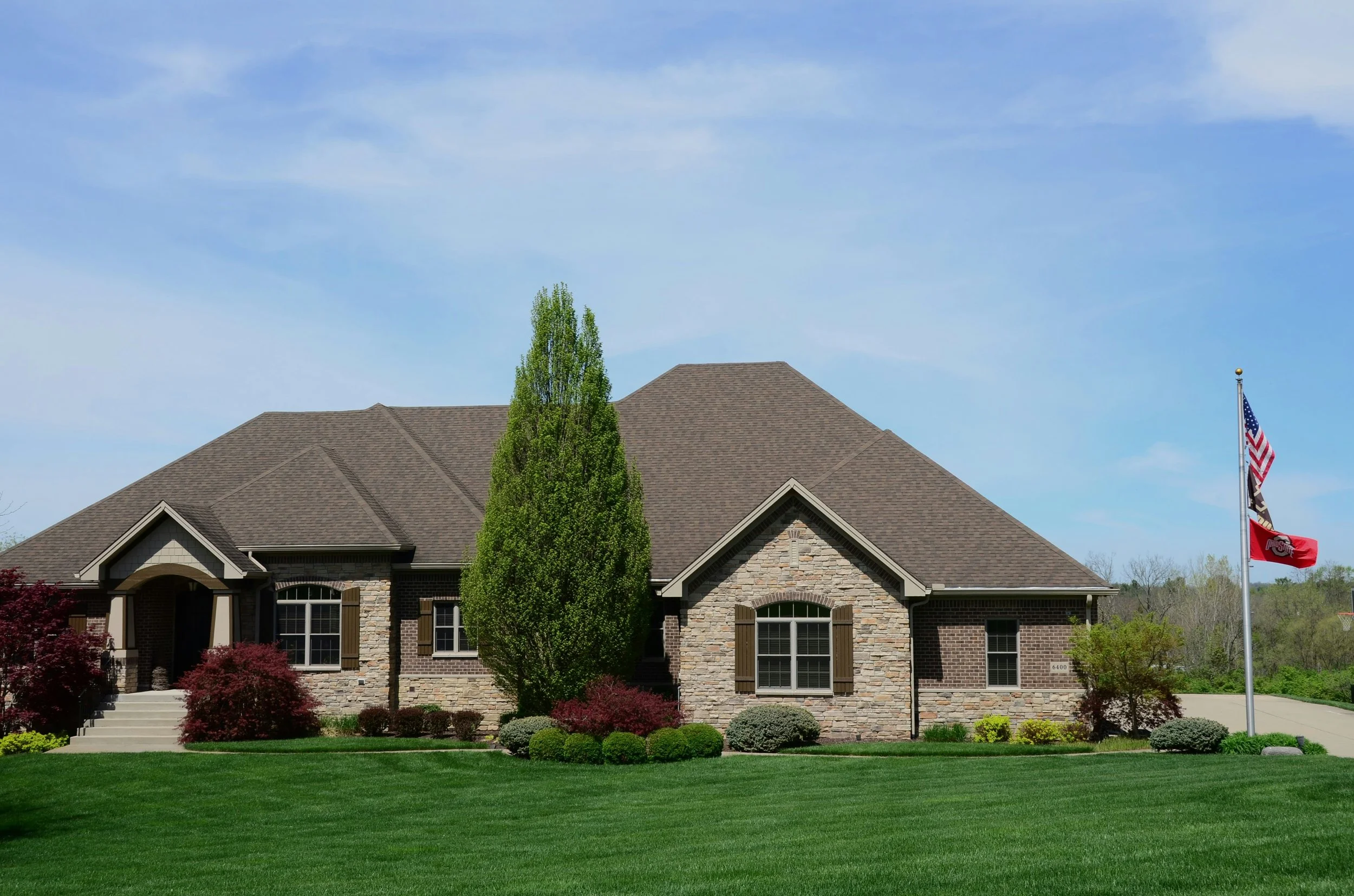 A suburban house with a brick and stone exterior, multiple windows with brown shutters, and a front porch, surrounded by a manicured lawn and bushes. There are two flags on poles to the right: an American flag and a red Ohio State flag. A large, tall
