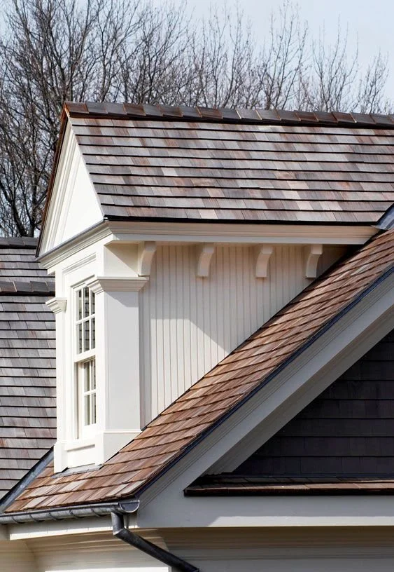 Close-up of house roof with brown shingles, dormer window with white trim, and leafless trees in background.