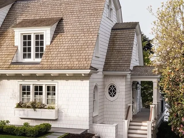 White house with brown shingle roof, dormer windows, front porch, and a small garden with bushes.