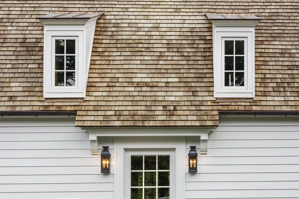 Front of a house with blonde shingle roof, two dormer windows, white siding, a central door with a window, and two black lantern-style exterior lights.
