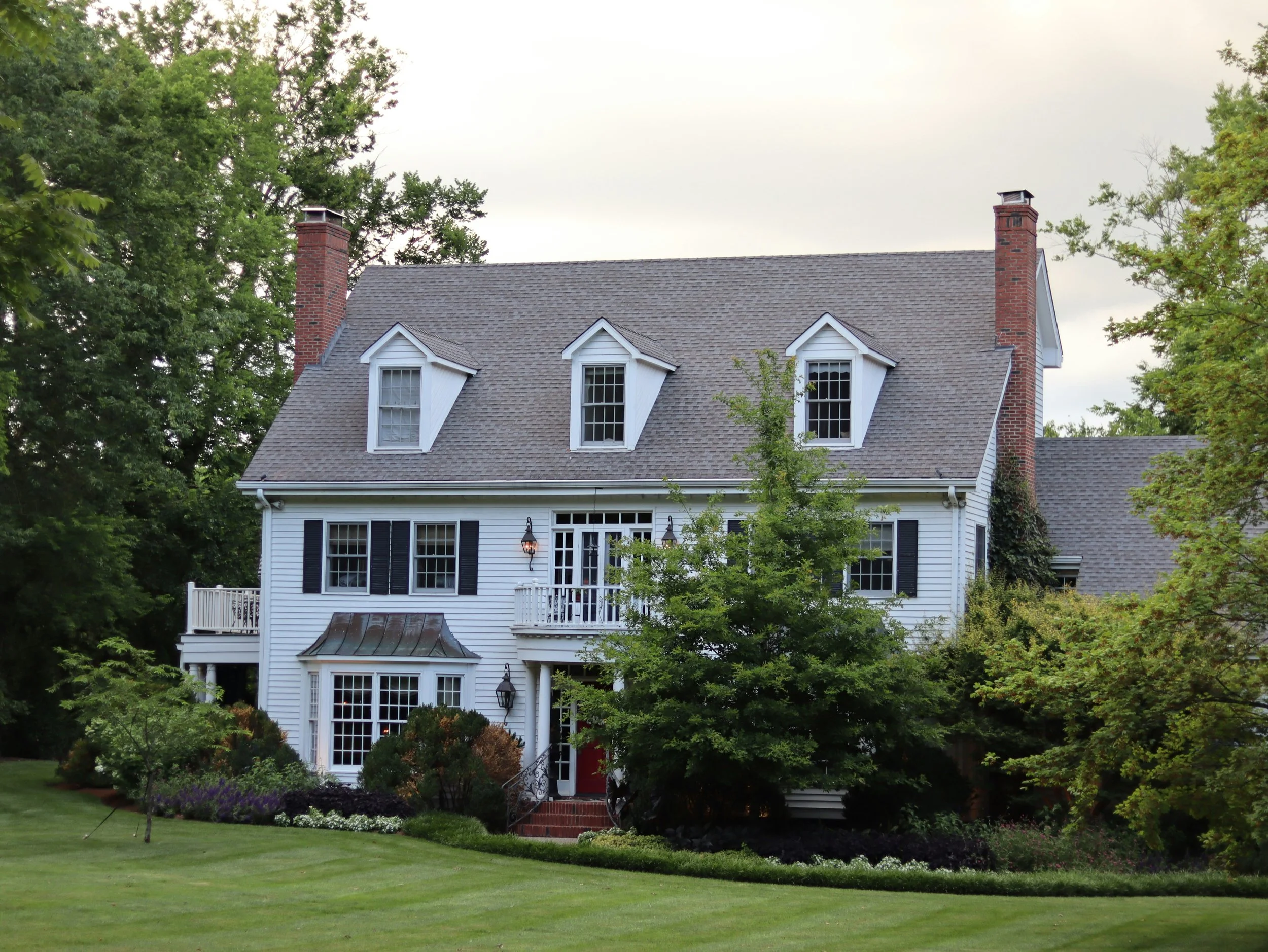 A large white house with three peaked dormer windows on the roof and a red front door, surrounded by green trees and plants, with a well-maintained lawn in the foreground.